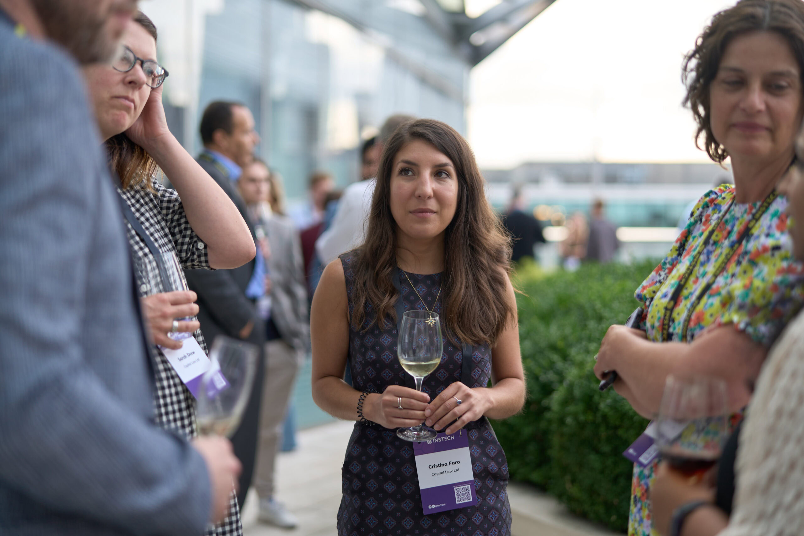 Alt text: Candid event photography by Adam Toth at Tadam Photography, capturing a young woman with long brown hair holding a wine glass at a professional networking event in London. The setting includes a blurred background of attendees, suggesting a vibrant outdoor gathering, possibly in East London. The image captures a natural, friendly atmosphere with soft, diffused