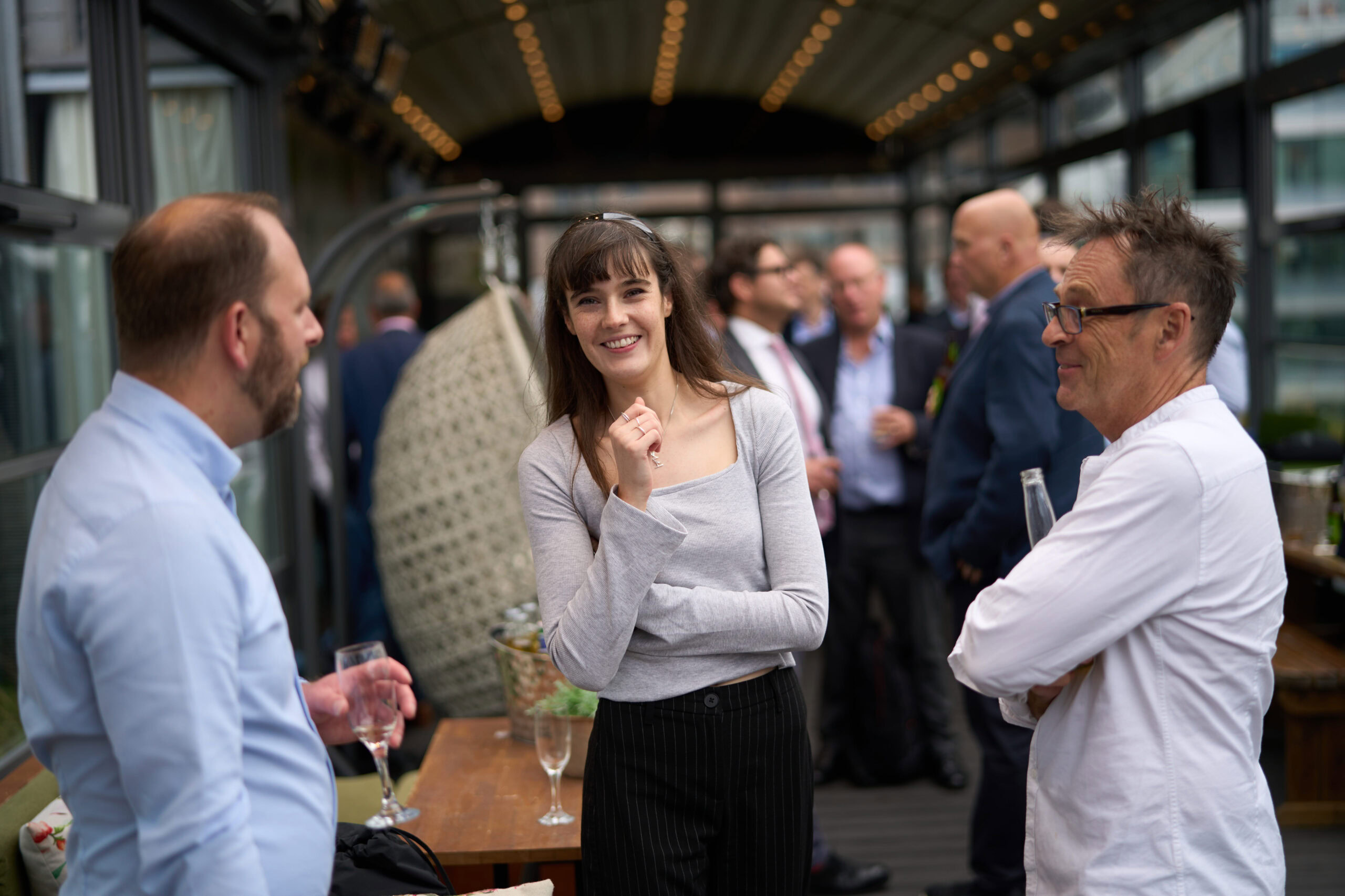 Alt text: Event photography by Adam Toth of Tadam Photography capturing a lively group interaction at a London venue. A smiling woman in a casual gray outfit engages confidently with two men in a relaxed, social setting. The composition uses natural lighting with a softly blurred background that enhances the candid and engaging atmosphere, typical of Tadam Photography's style,