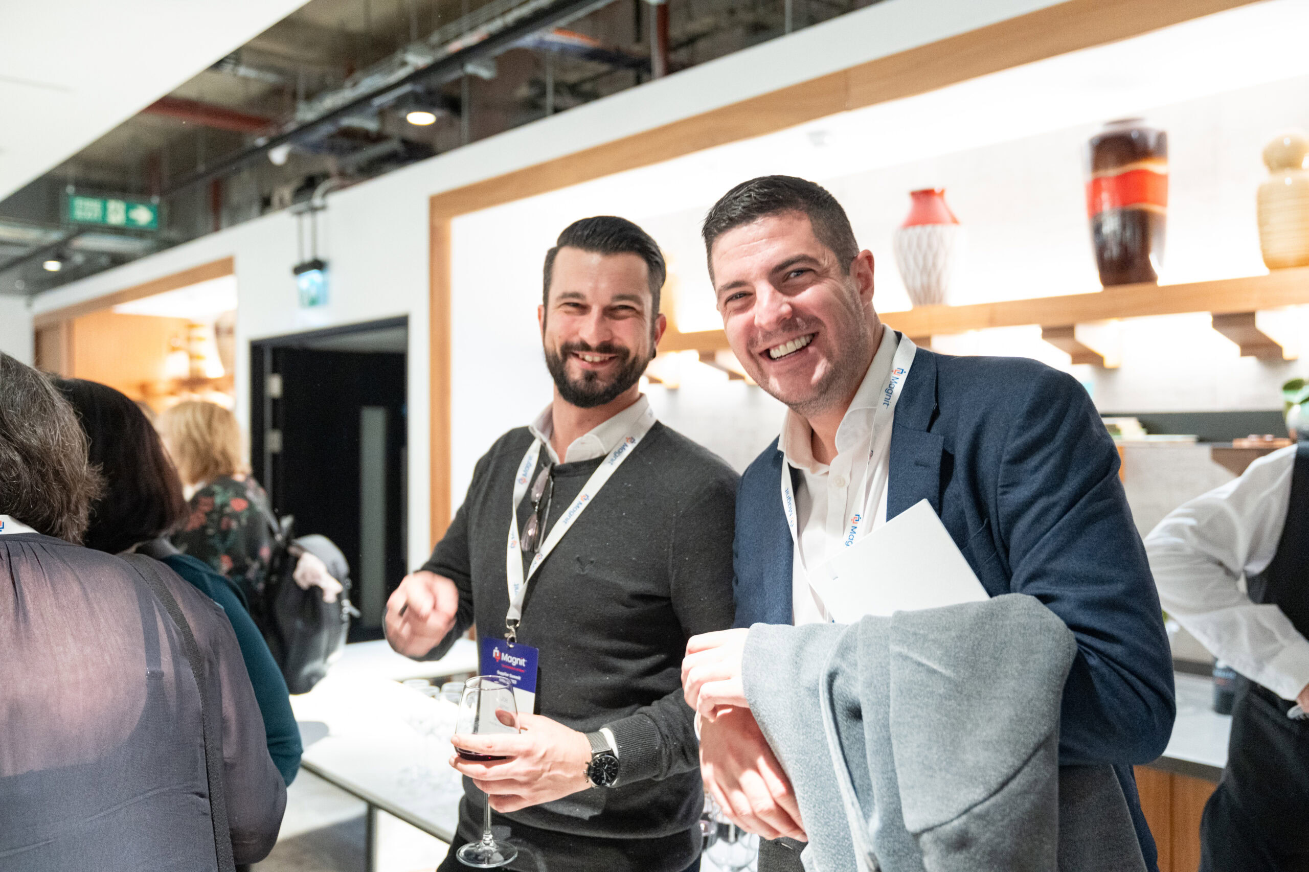 A lively event photography shot by Adam Toth at Tadam Photography, capturing two smiling professionals enjoying a corporate gathering in London. They display a natural rapport, dressed in smart casual attire with name badges, set against the backdrop of a modern office space. The clean composition and well-utilized ambient lighting create an inviting and authentic mood, highlighting Toth's