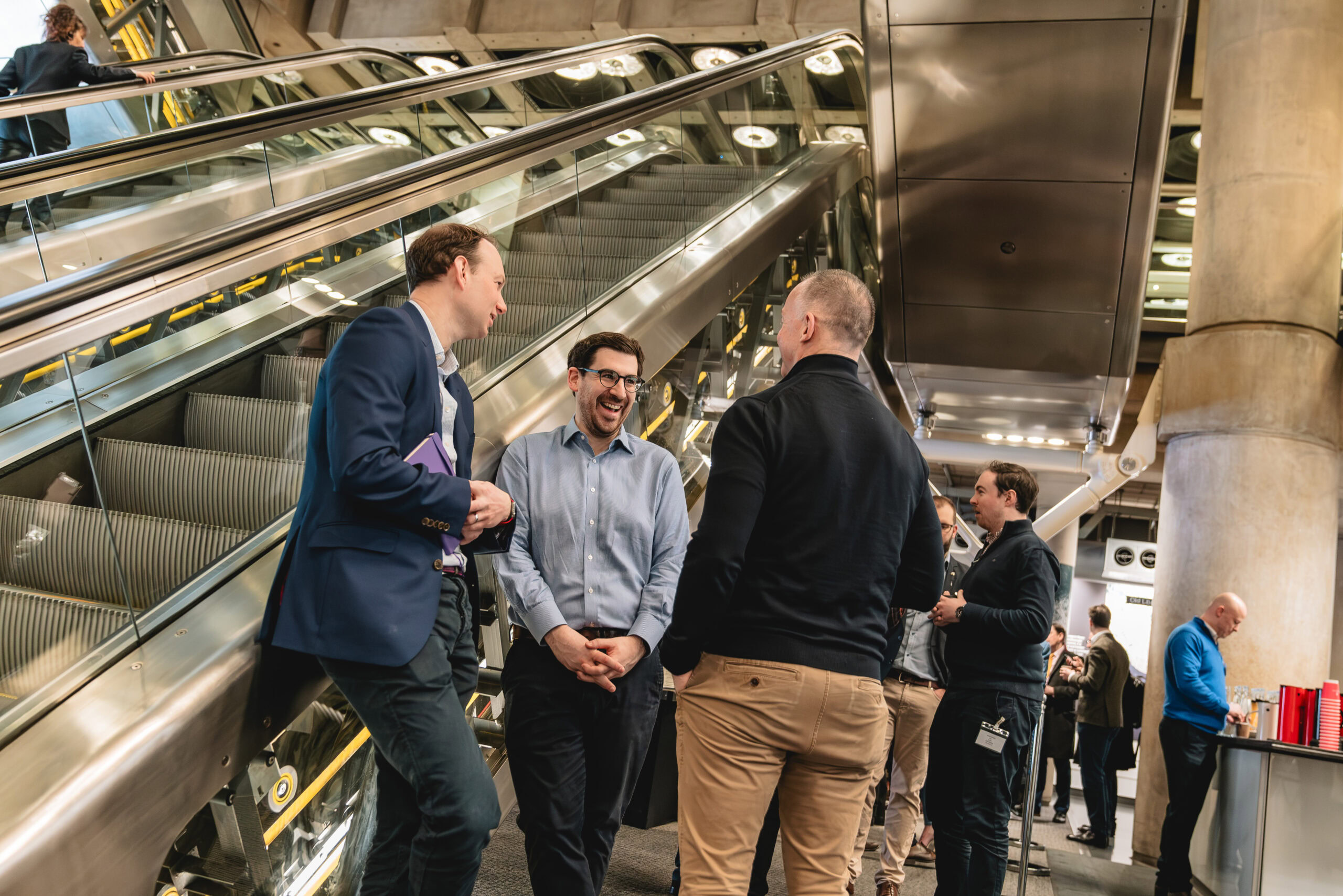 Professional networking event photography capturing a group of men engaged in a lively conversation at a modern London venue. The spontaneous and relaxed poses create an authentic atmosphere, enhanced by the stylish urban setting near an escalator. The image features dynamic composition with natural lighting, skillfully captured by Adam Toth for Tadam Photography, highlighting vibrant corporate interactions in East London.
