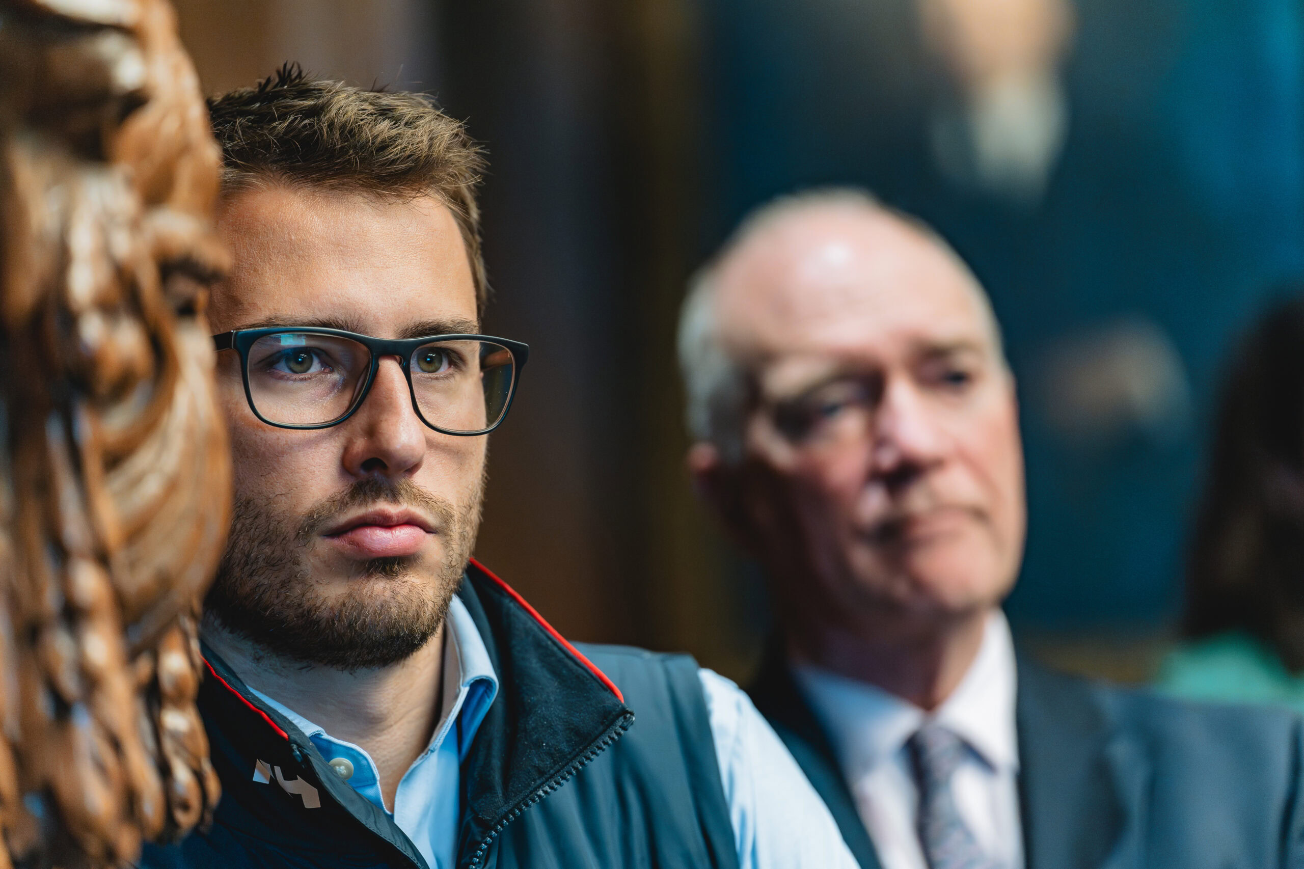 Corporate headshot by Adam Toth of Tadam Photography features a focused man with glasses in a smart-casual outfit, set against a blurred background during an event. Captured with sharp, professional lighting that emphasizes his determined expression, this image evokes a serious, professional mood. Photographed in London, it showcases Toth's expertise in dynamic event