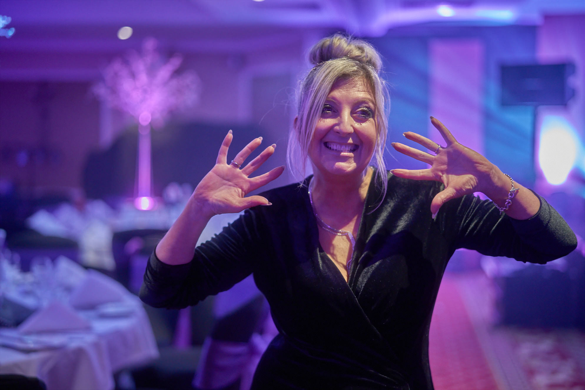 Woman smiling and posing with hands raised at an indoor event with purple lighting.