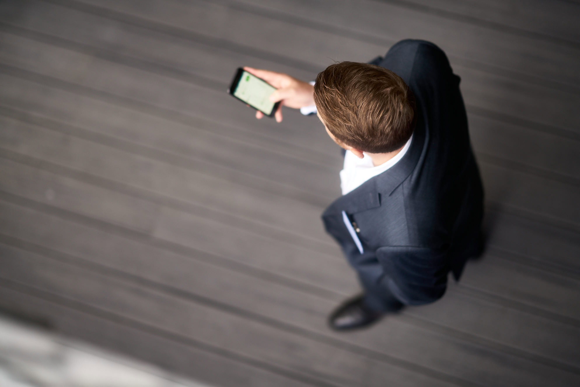 A businessman in a suit looking at his smartphone while standing on wooden flooring.