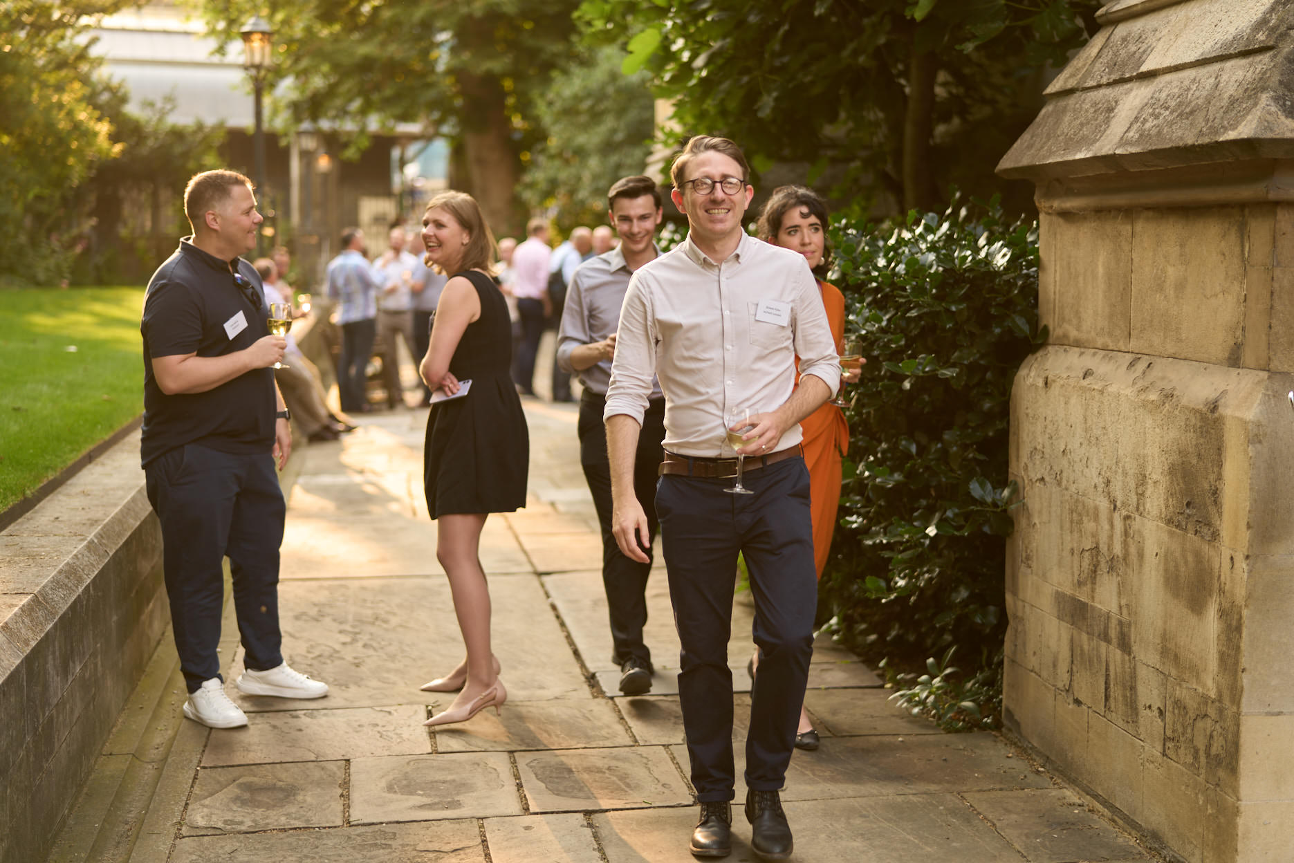 A group of individuals socialising outdoors, some holding drinks, in a garden setting.