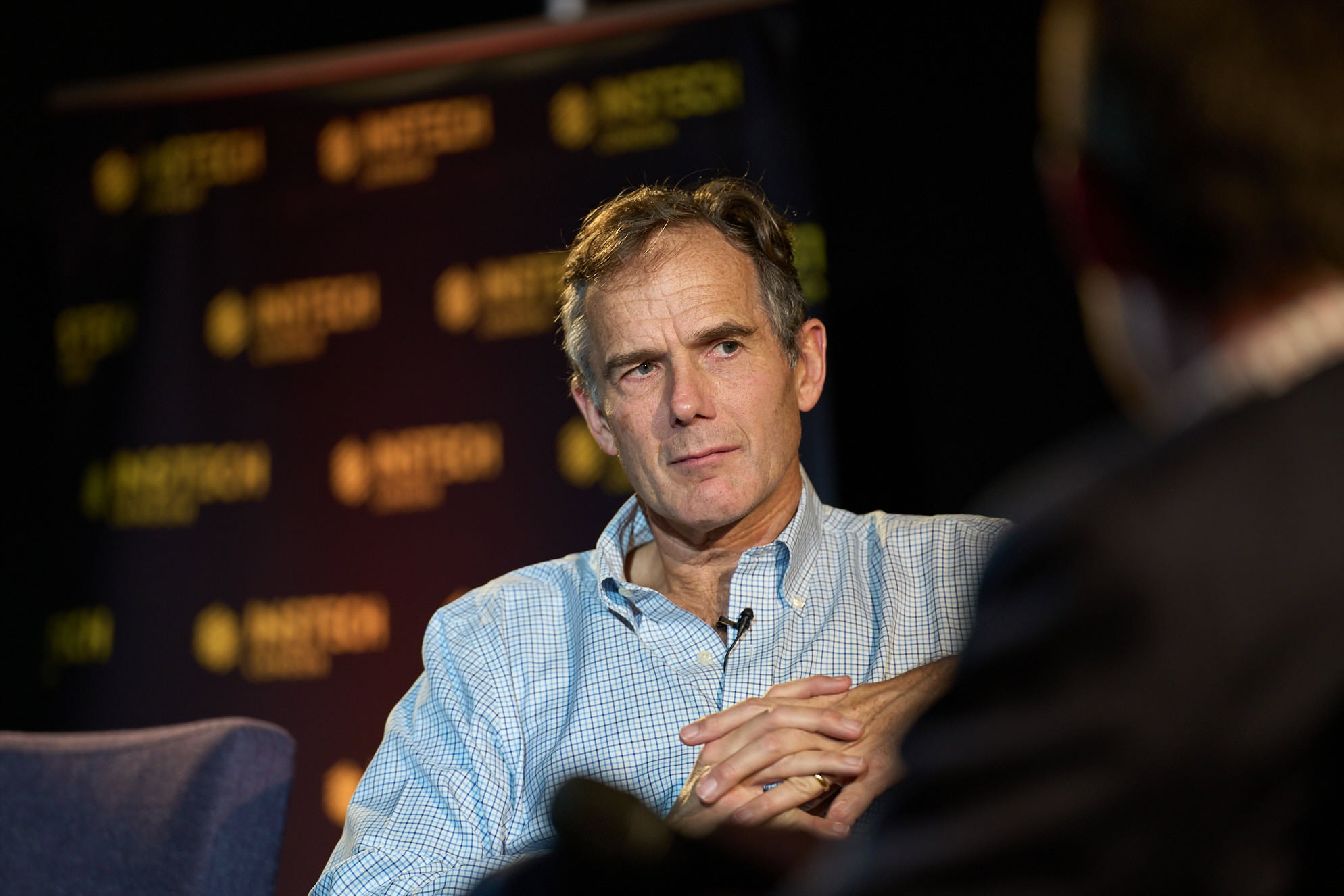 Man wearing a blue checkered shirt sitting during an interview with a blurred background.