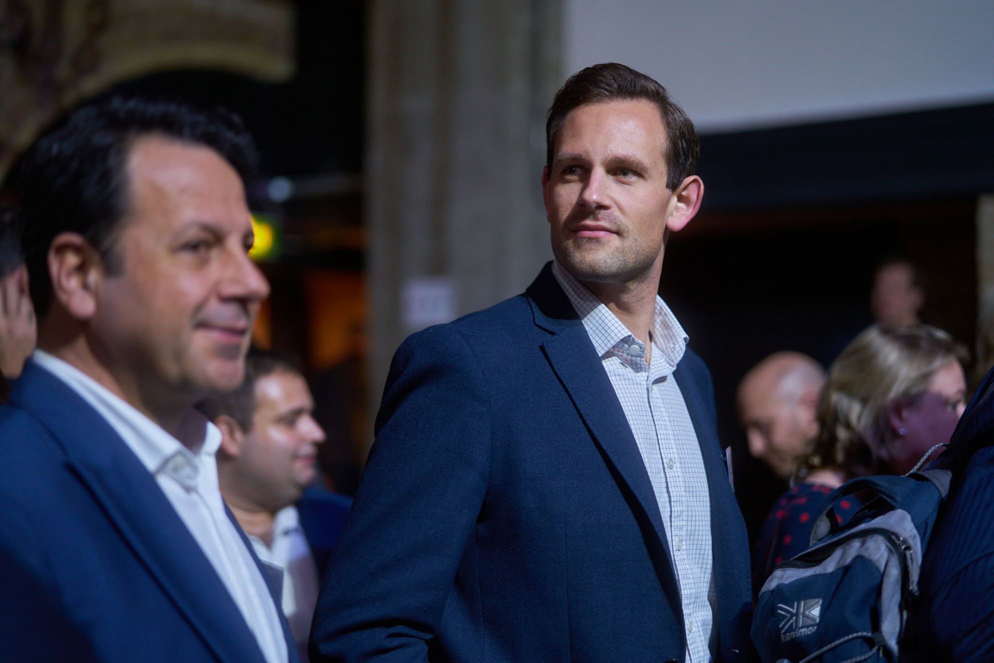 Two men in business attire standing together at an event, one looking away from the camera.