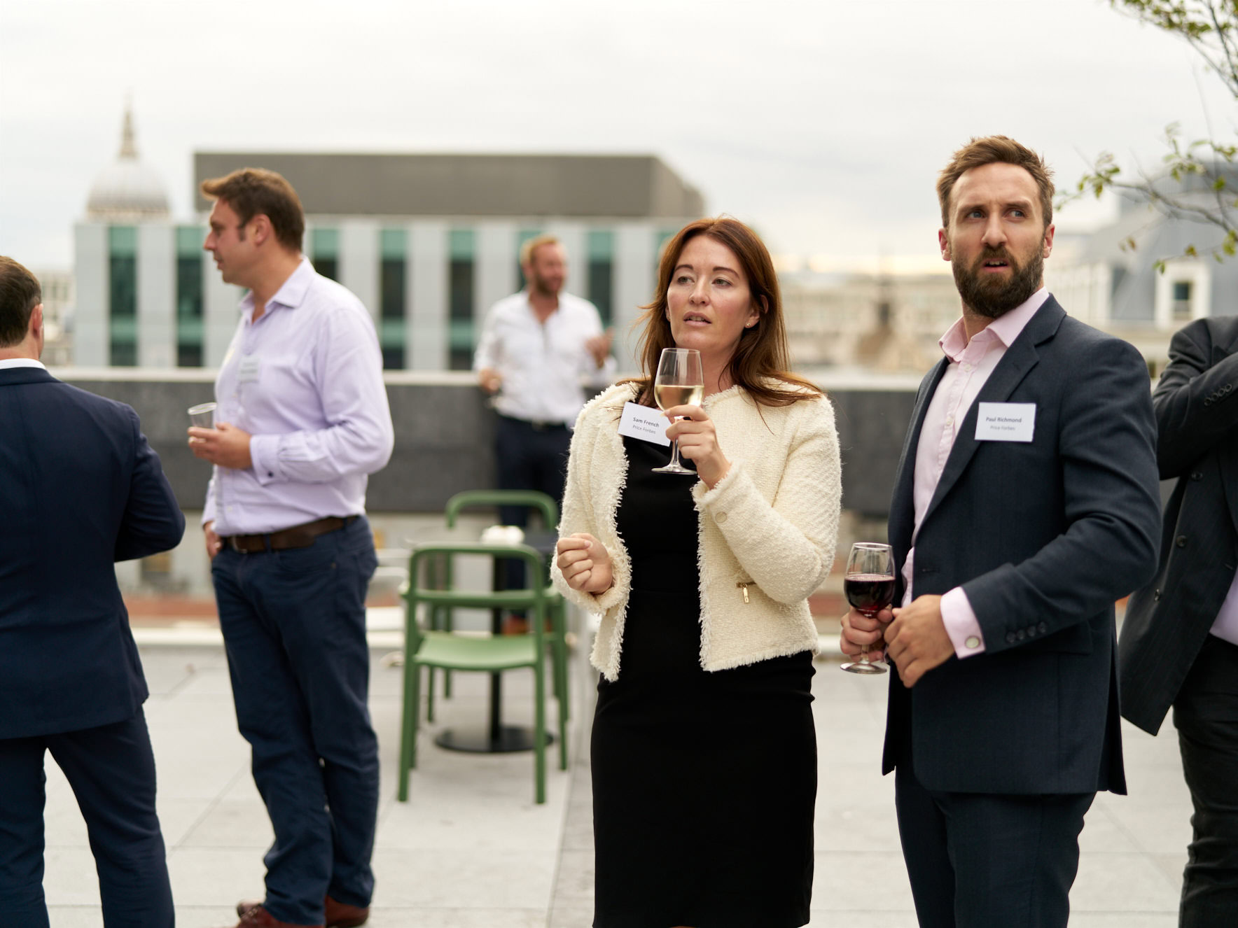 Two professionals holding glasses of wine at a rooftop networking event with others in the background.