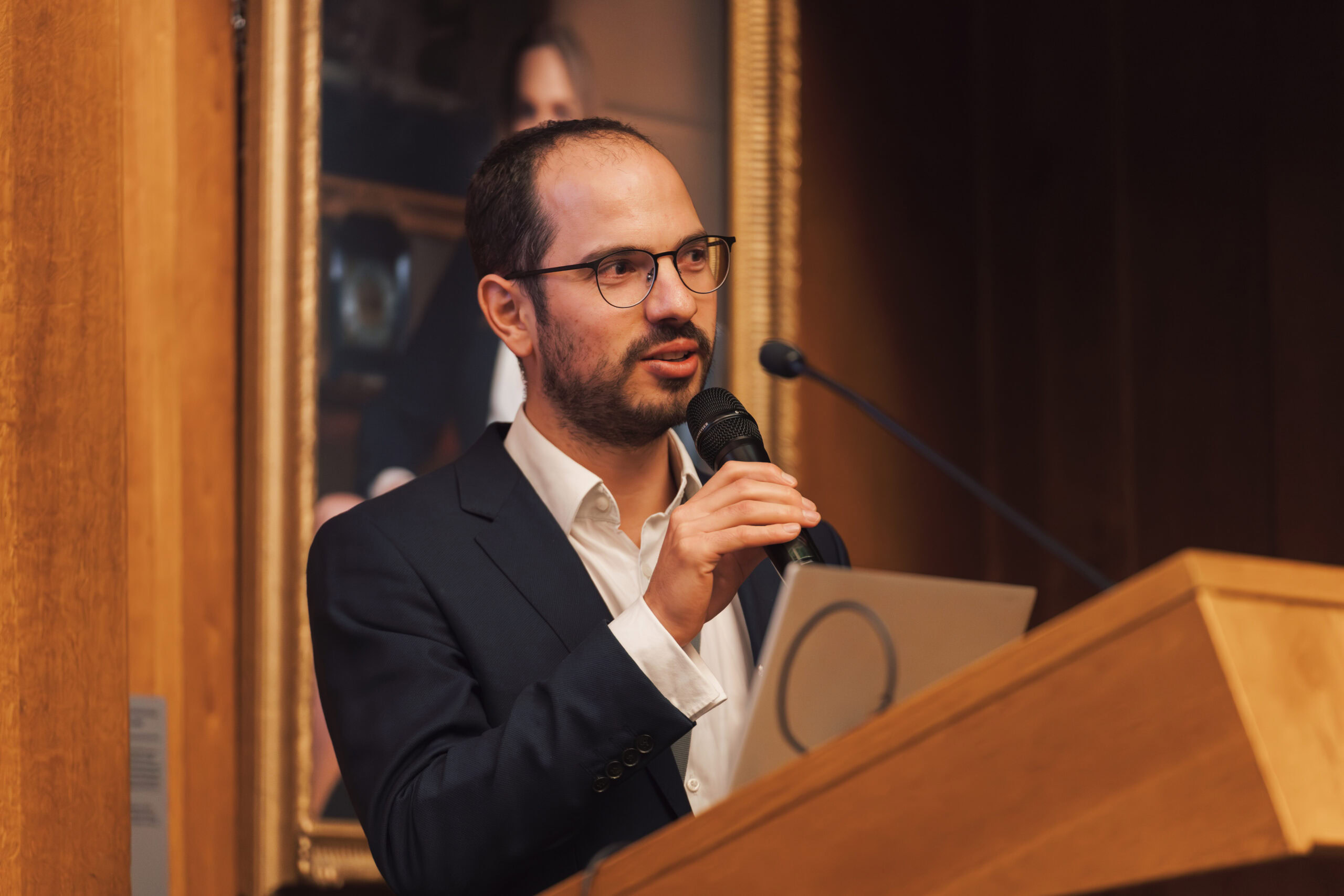 A man in a suit speaking into a microphone at a podium during an event.