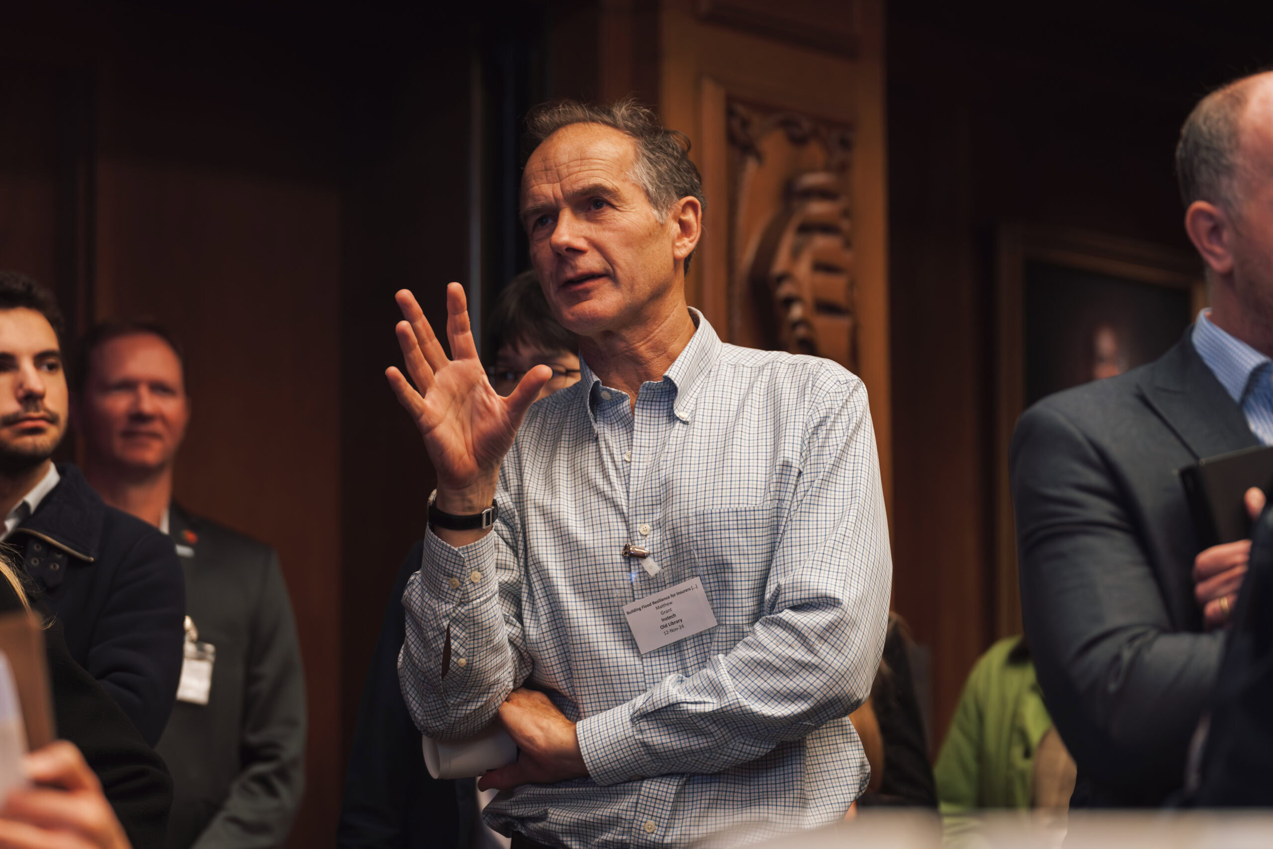 A man in a checked shirt gesturing while speaking at a conference with an audience in the background.
