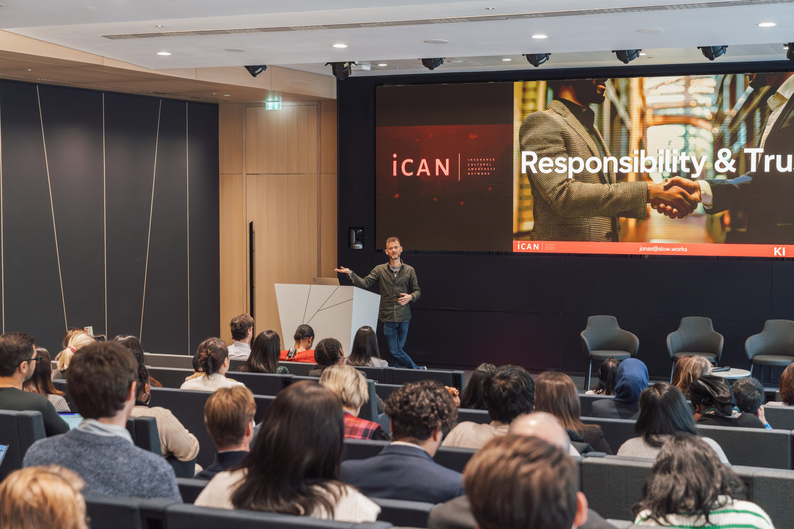 A speaker presenting in front of an audience at a conference with a screen displaying 'Responsibility & Trust'.