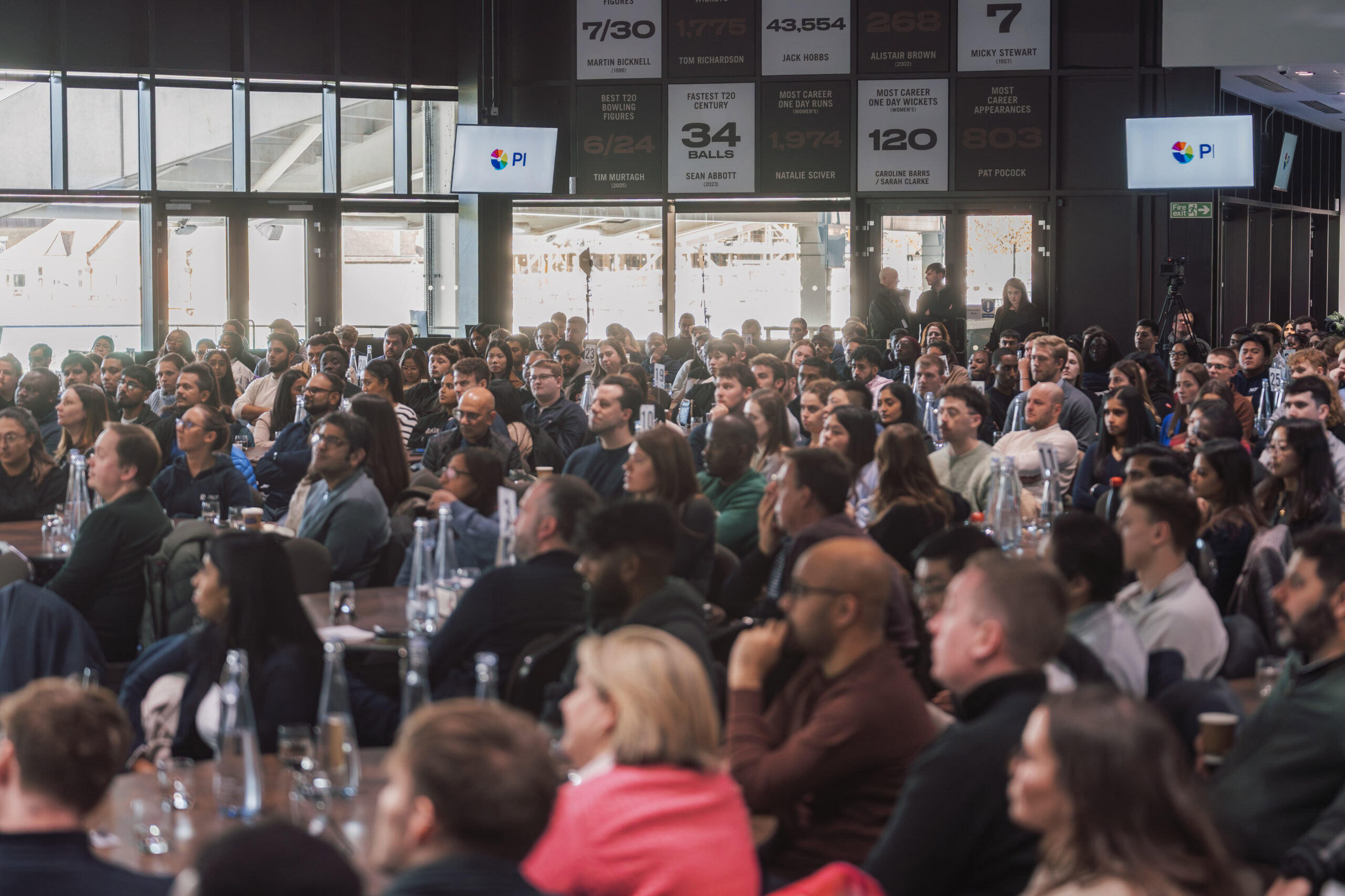 Large audience seated at tables during a conference event with screens displaying information.