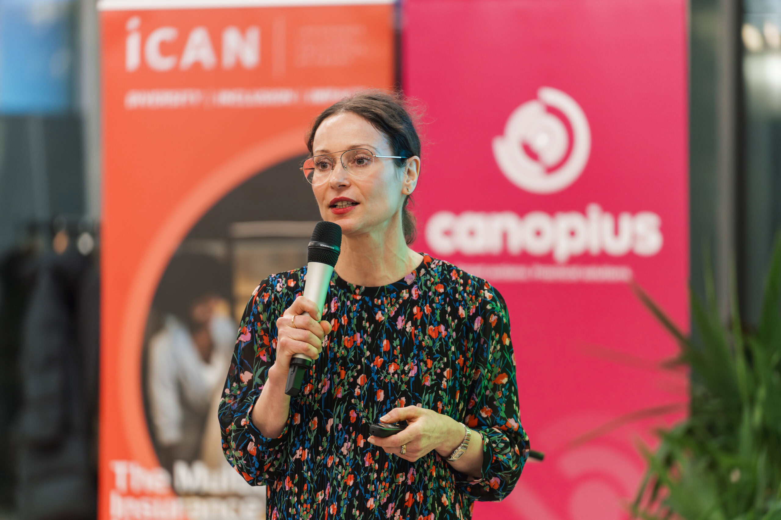 A woman with glasses speaking into a microphone at a conference event with banners in the background.