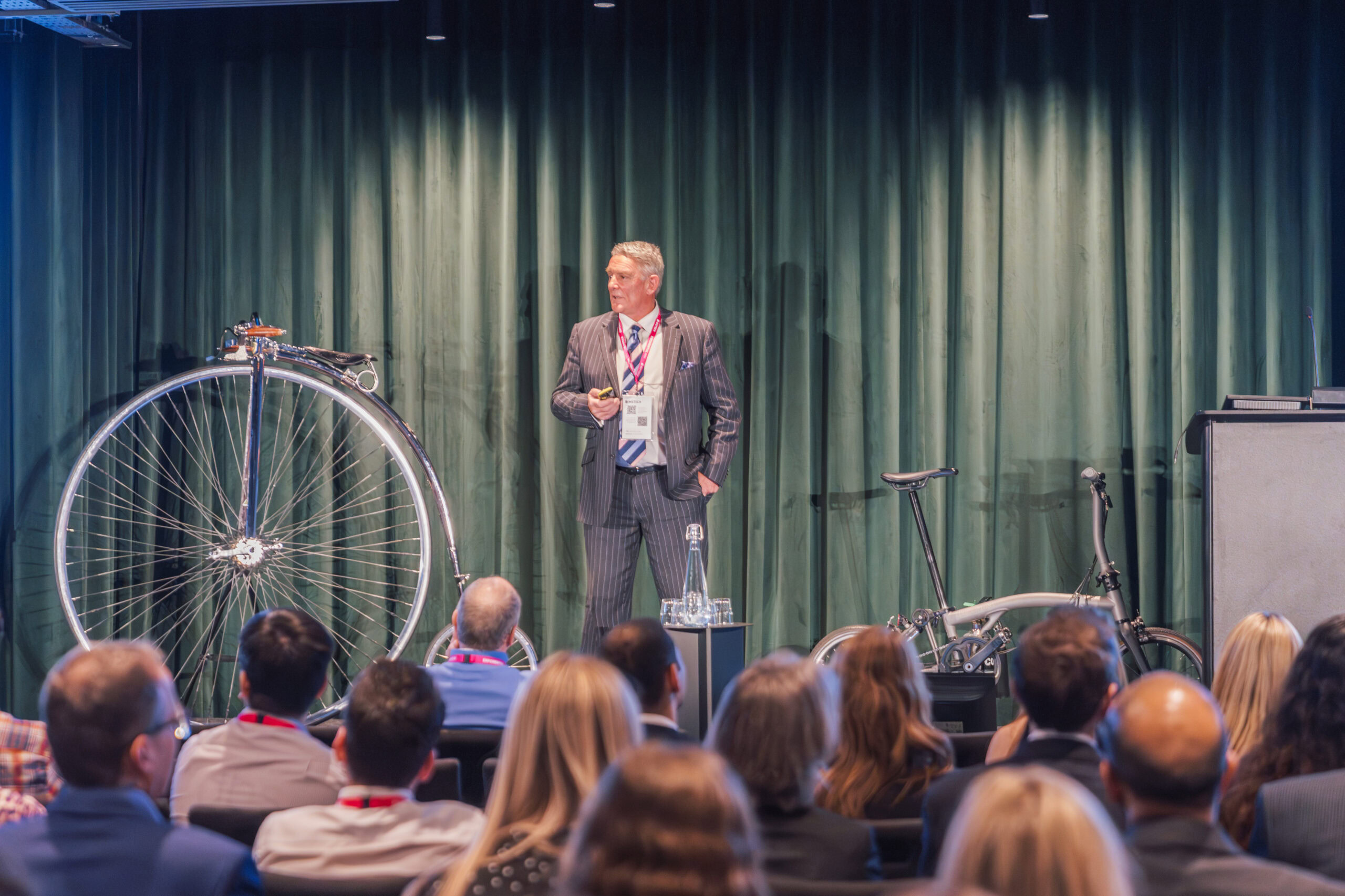 Speaker presenting in front of a vintage penny-farthing bicycle and a modern bicycle at a conference.