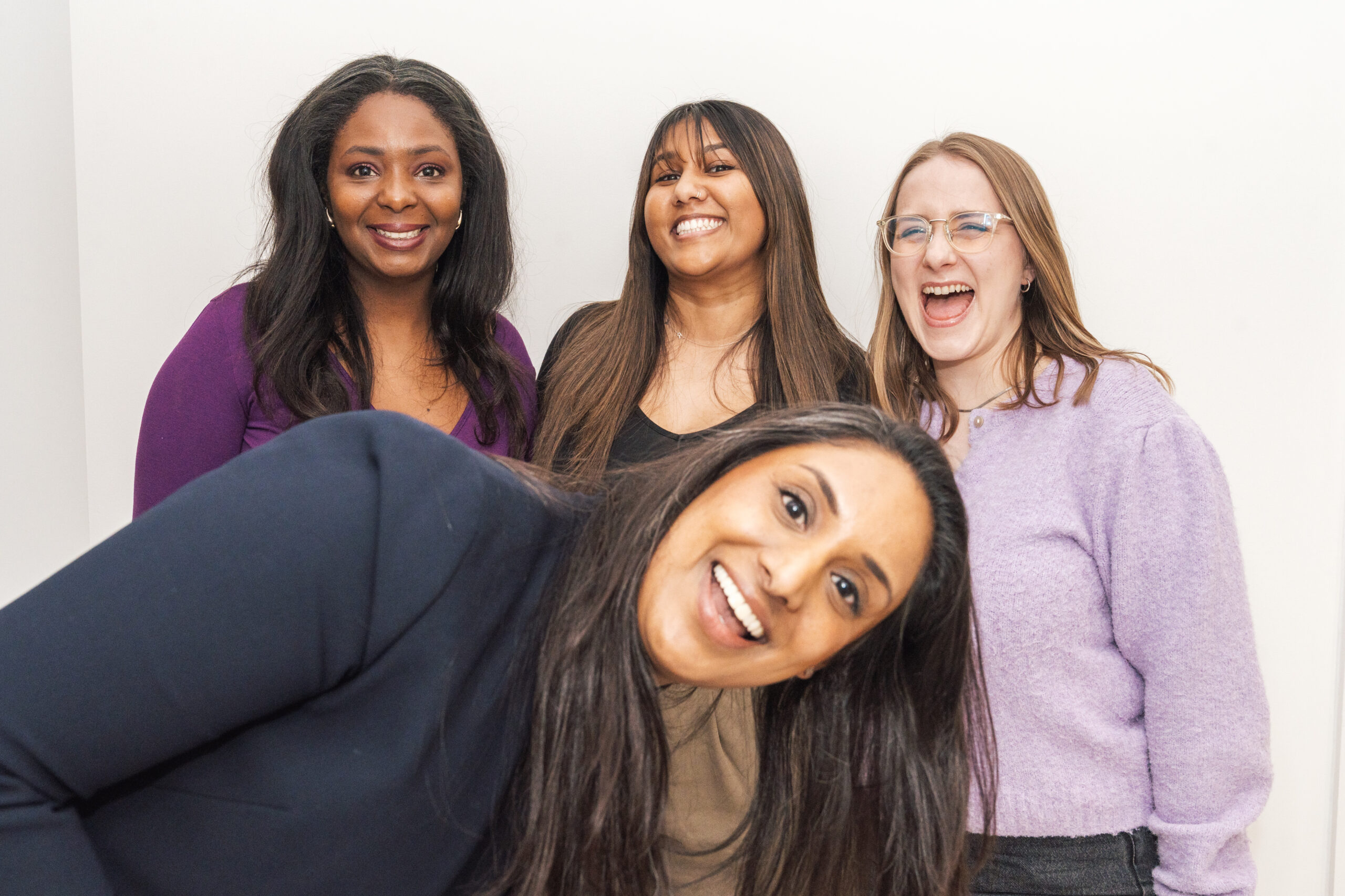 Four women smiling and posing together in a relaxed indoor setting, with one woman leaning forward.