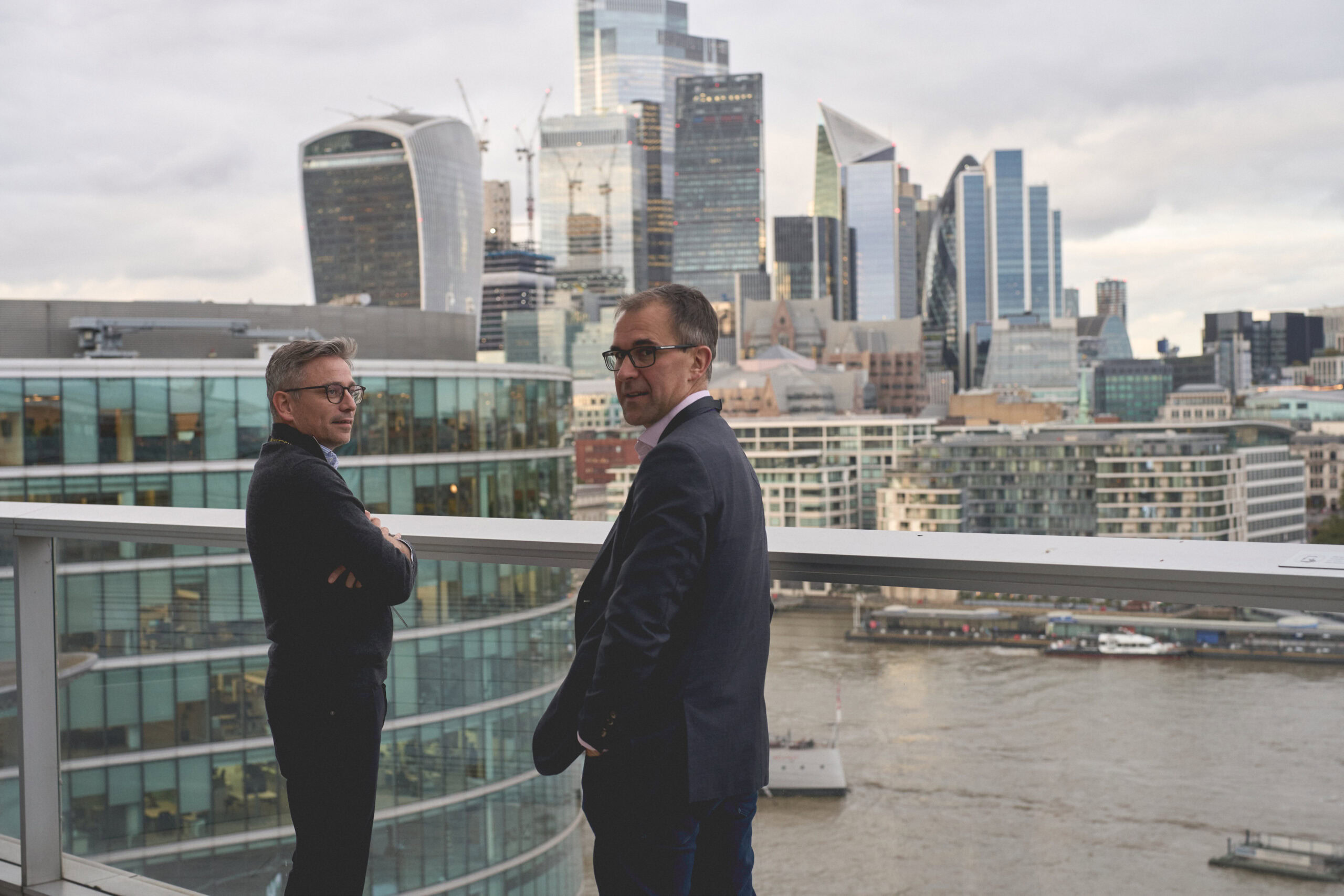 Two men standing on a balcony overlooking the London skyline with modern buildings and the River Thames.