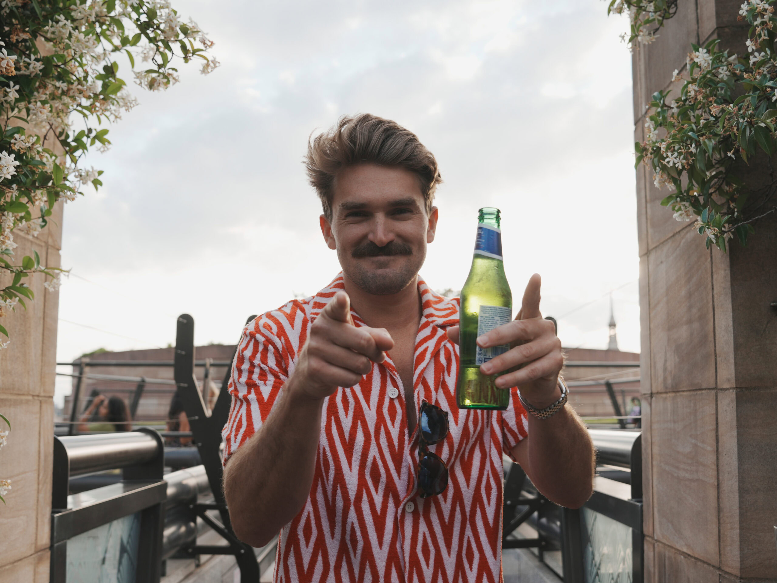 Man in a patterned shirt holding a green bottle of beer and pointing at the camera.