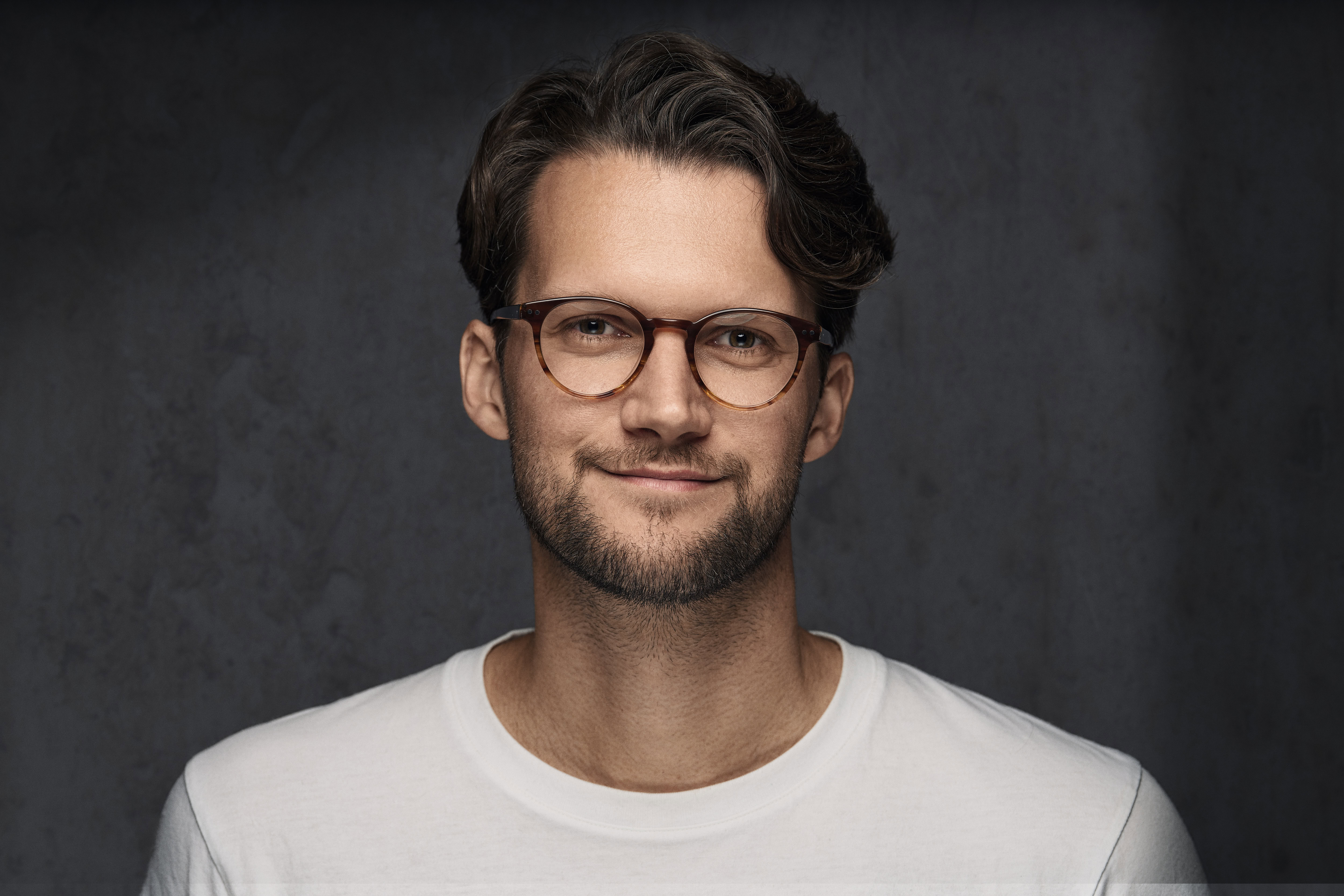 Corporate headshot of a man with glasses in a relaxed pose, wearing a white t-shirt. Captured by Adam Toth of Tadam Photography in London, this image uses professional studio lighting to create a soft, natural atmosphere against a dark, clean background.