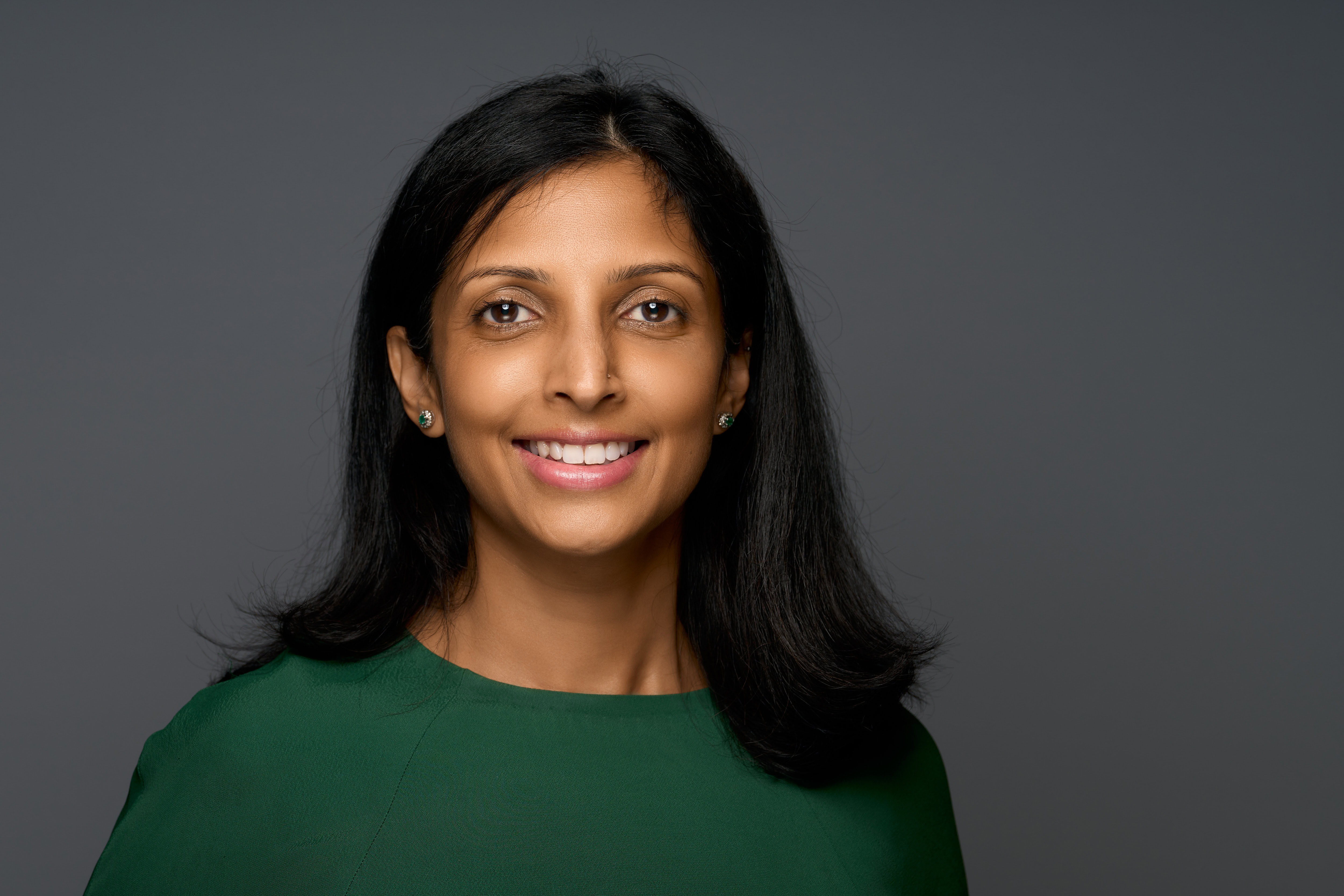 Corporate headshot of a woman with long dark hair and a green top, captured by Adam Toth of Tadam Photography. The subject displays a confident smile in a natural pose against a neutral backdrop. Professional studio lighting creates a balanced and polished look. Shot in London, enhancing the urban professional atmosphere.
