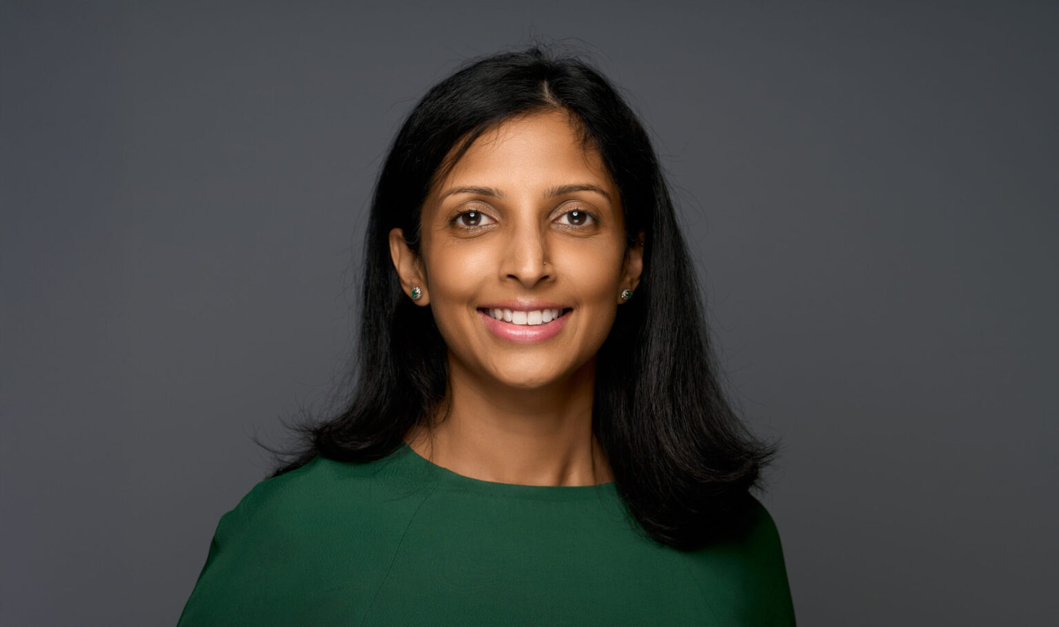 Harsitha smiling brightly in a dark green top, long dark hair, on a soft grey studio backdrop
