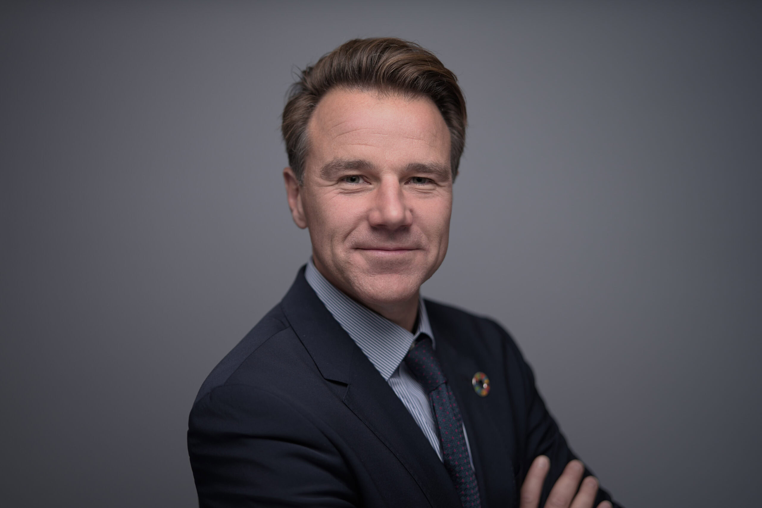 Man with swept-back grey-blond hair in a navy suit and tie with a pin badge, arms crossed, confident half-smile