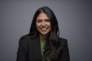 Woman with long dark wavy hair in a black blazer over a green top, laughing warmly on a grey backdrop