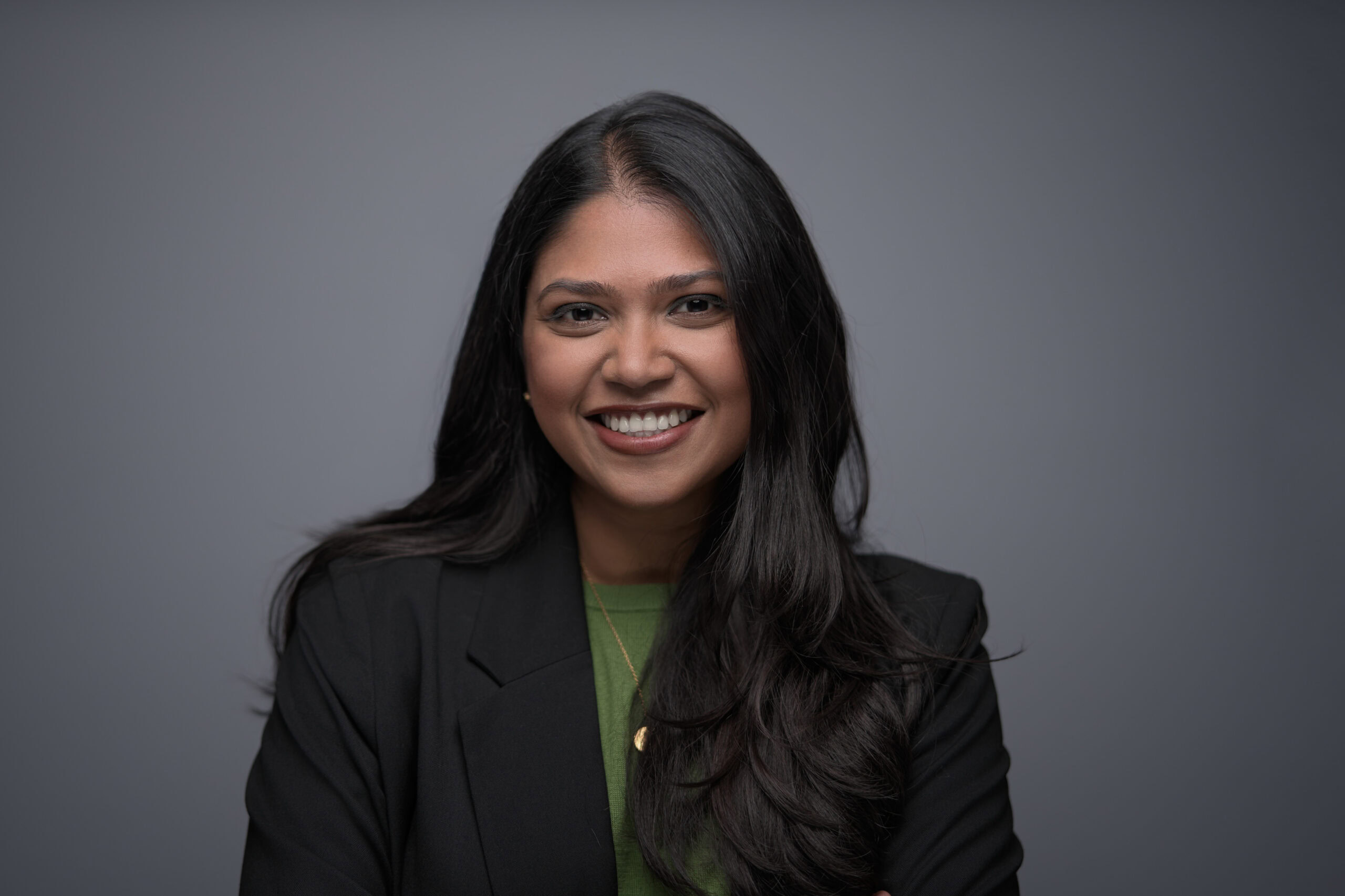 Woman with long dark wavy hair in a black blazer over a green top, laughing warmly on a grey backdrop