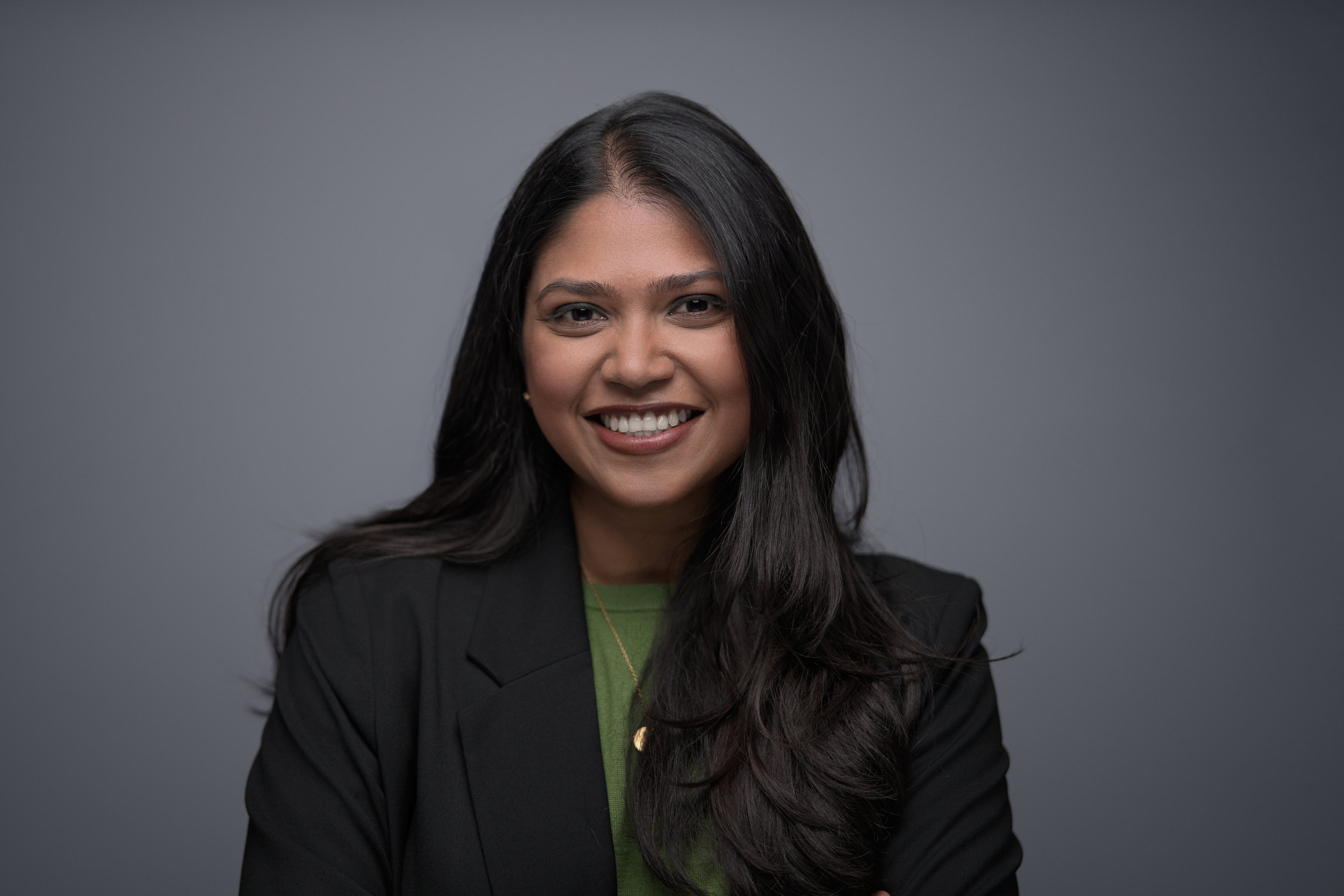 Corporate headshot of a smiling professional woman in a black blazer against a clean, gray backdrop. The natural pose and soft studio lighting create an approachable and confident appearance. Captured by Adam Toth at Tadam Photography, this image reflects a modern and polished style typical of business portraits in London.