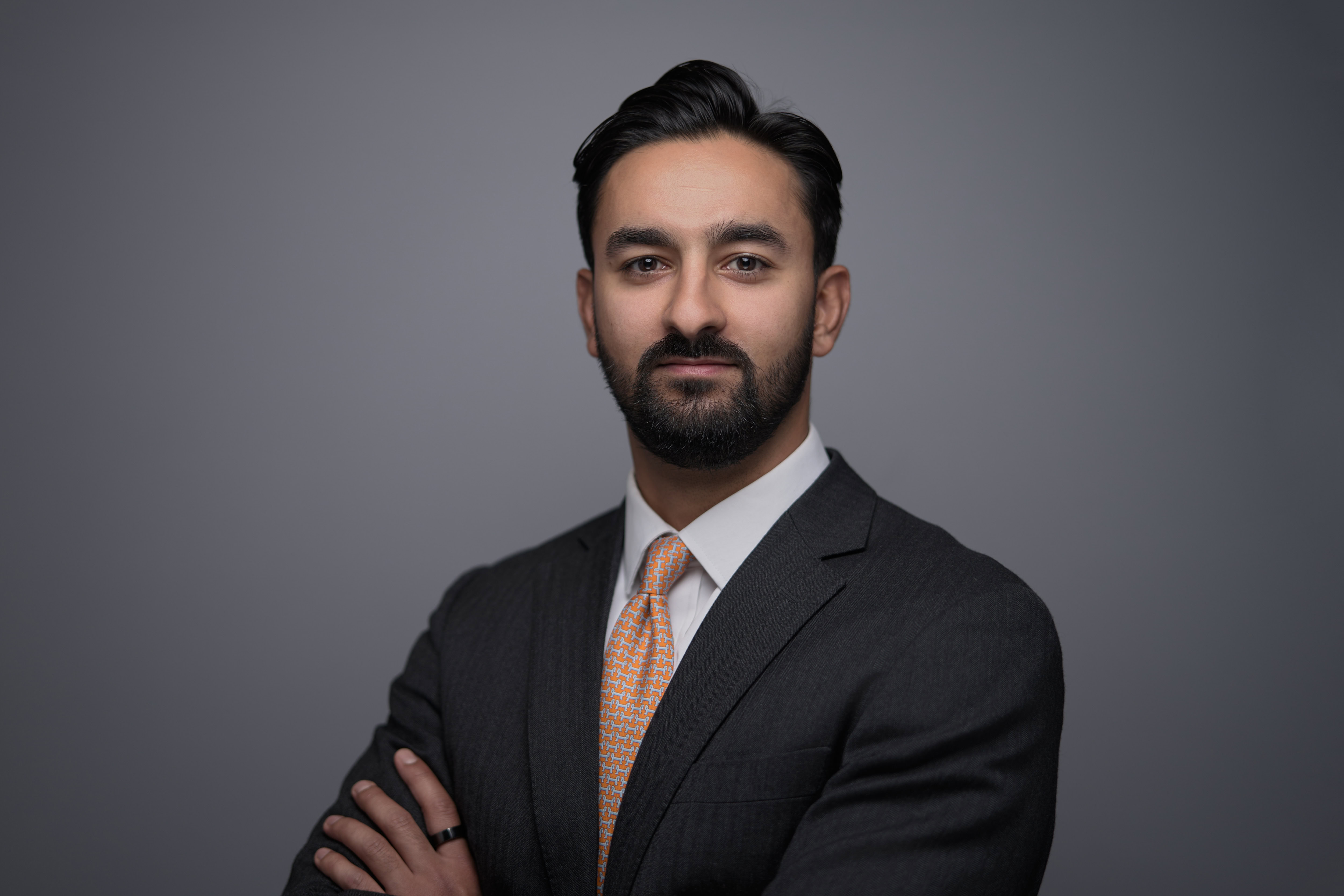 Corporate headshot featuring a confident professional in a tailored dark suit and checkered tie, arms crossed, set against a clean, grey background. The image captures a composed demeanor with professional studio lighting, highlighting an authentic expression. Photographed by Adam Toth of Tadam Photography in London, focusing on premium corporate imagery.