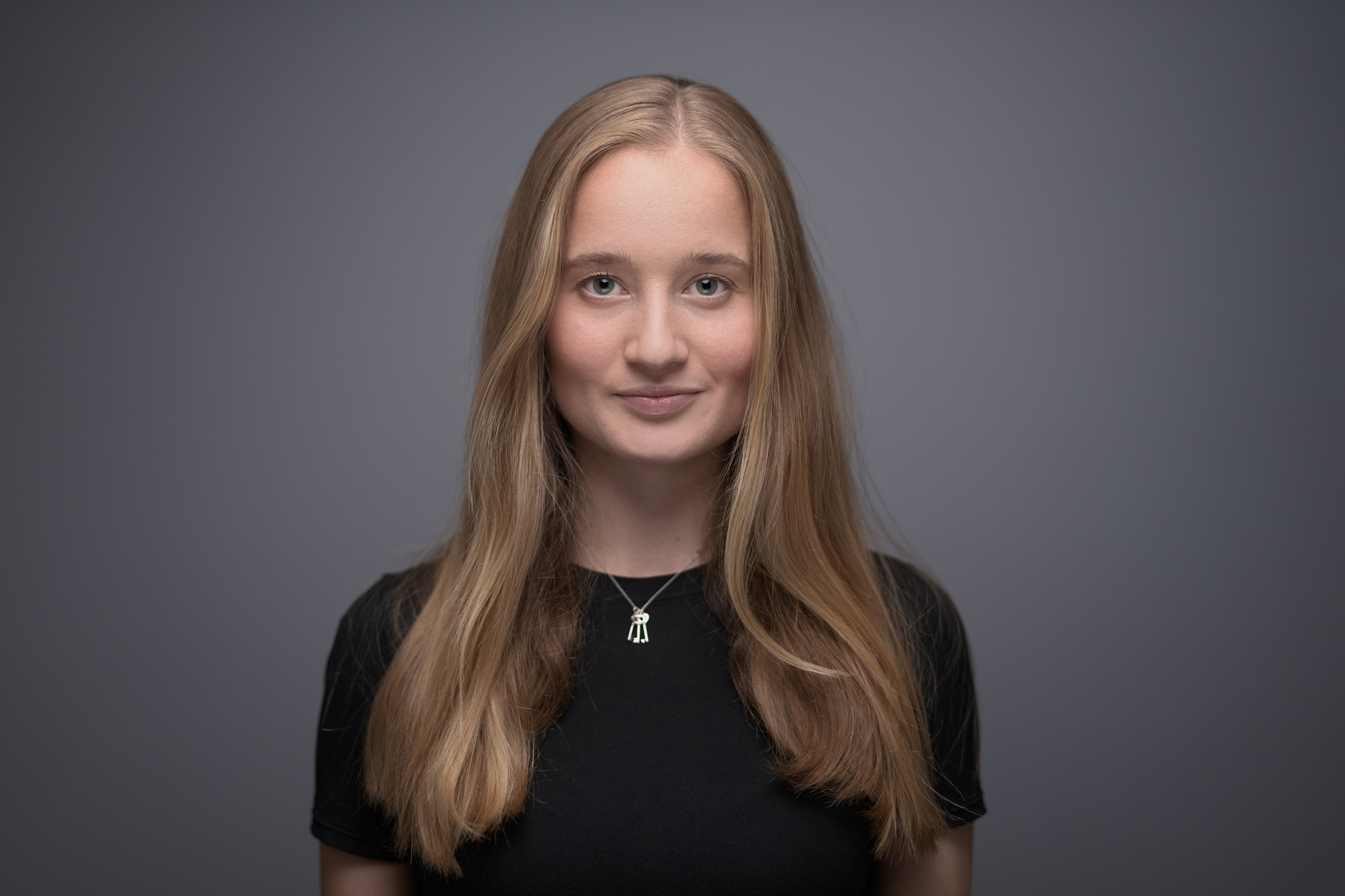 Corporate headshot of a young woman with long blonde hair, captured by Adam Toth at Tadam Photography in London. The subject stands against a soft, grey background, wearing a simple black top. Her expression is calm and confident, complemented by the professional studio lighting that highlights her natural features. This image exemplifies Adam Toth’s expertise in creating