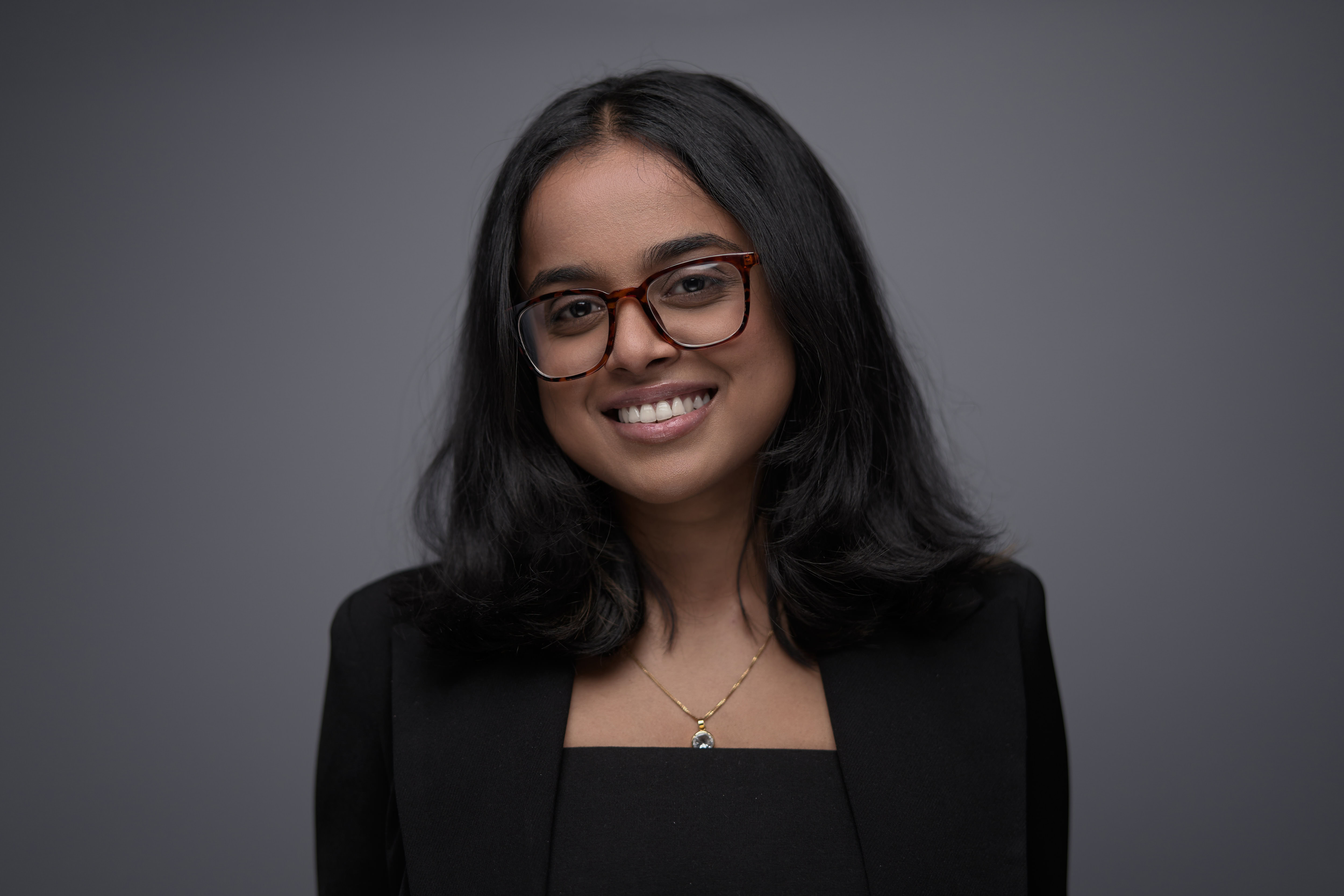 Corporate headshot photography by Adam Toth of Tadam Photography, featuring a confident professional woman with long black hair and stylish glasses, posed naturally in a tailored black outfit against a clean, gray background. The image utilizes professional studio lighting to highlight an authentic expression, creating a polished and approachable appearance. Captured in London, this portrait exemplifies the expertise