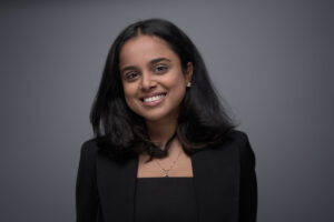 Woman with shoulder-length dark hair in a black blazer and delicate necklace, smiling brightly