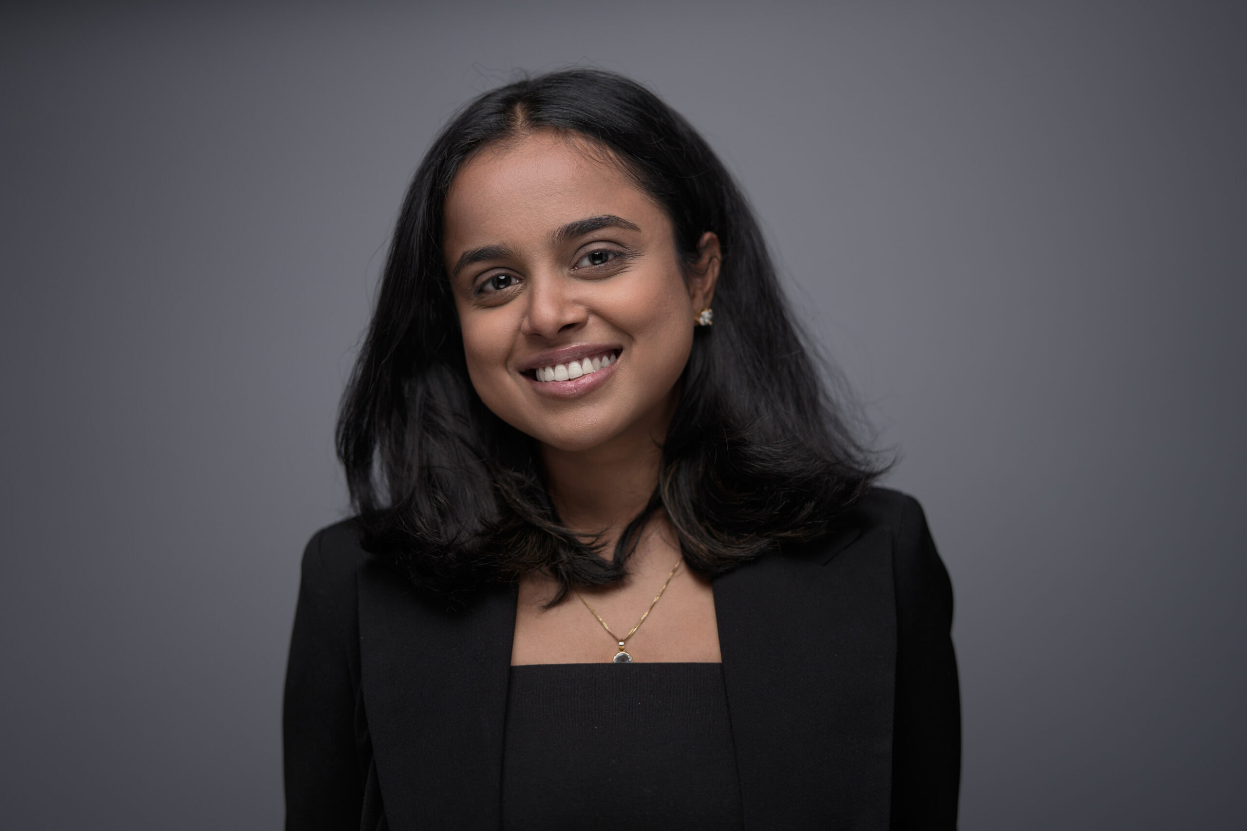 Woman with shoulder-length dark hair in a black blazer and delicate necklace, smiling brightly