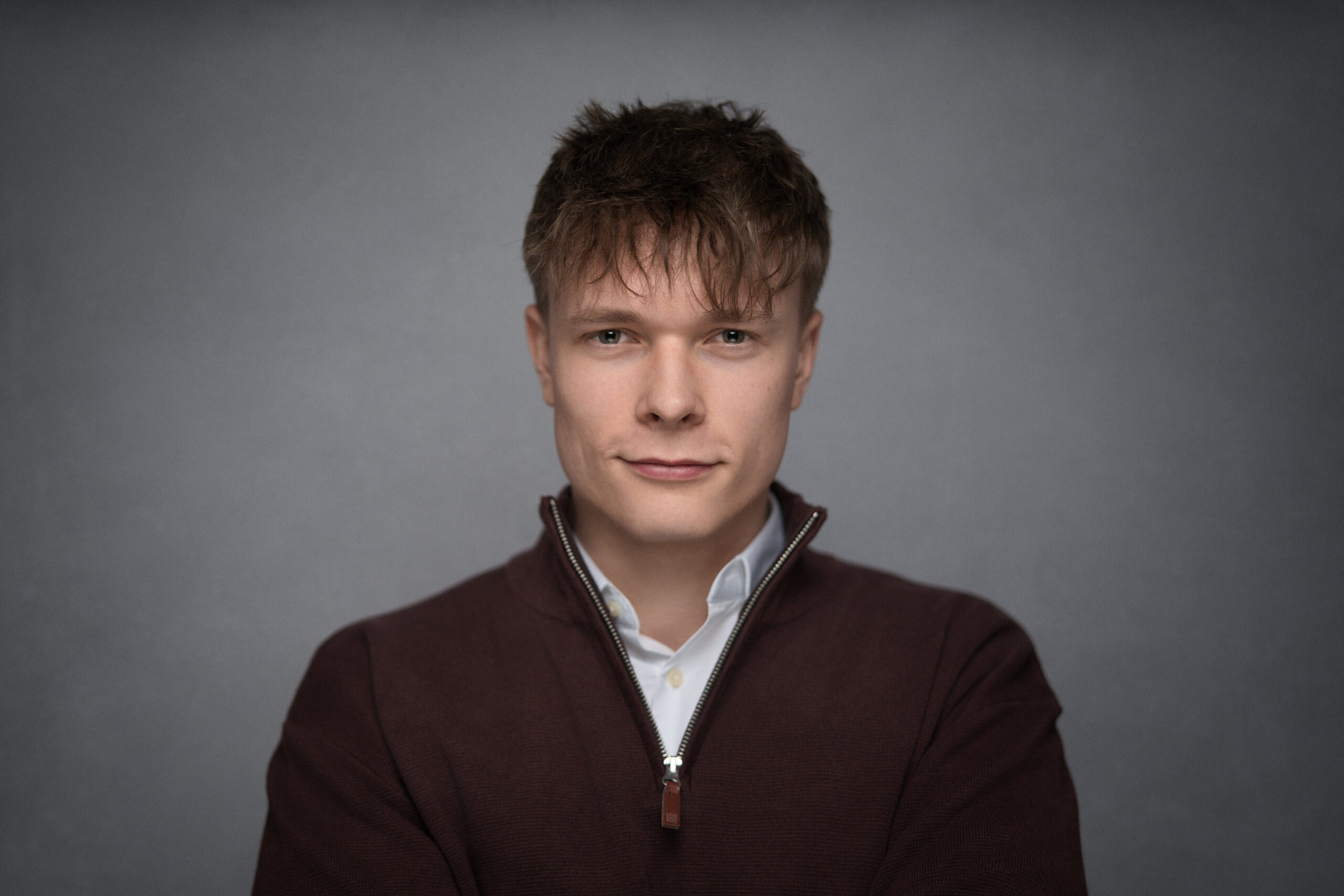 Young man with short tousled hair wearing a dark zip-up sweater over a white shirt on gray background