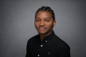 Man with braided hair in a black polo shirt, direct look, on a grey studio backdrop