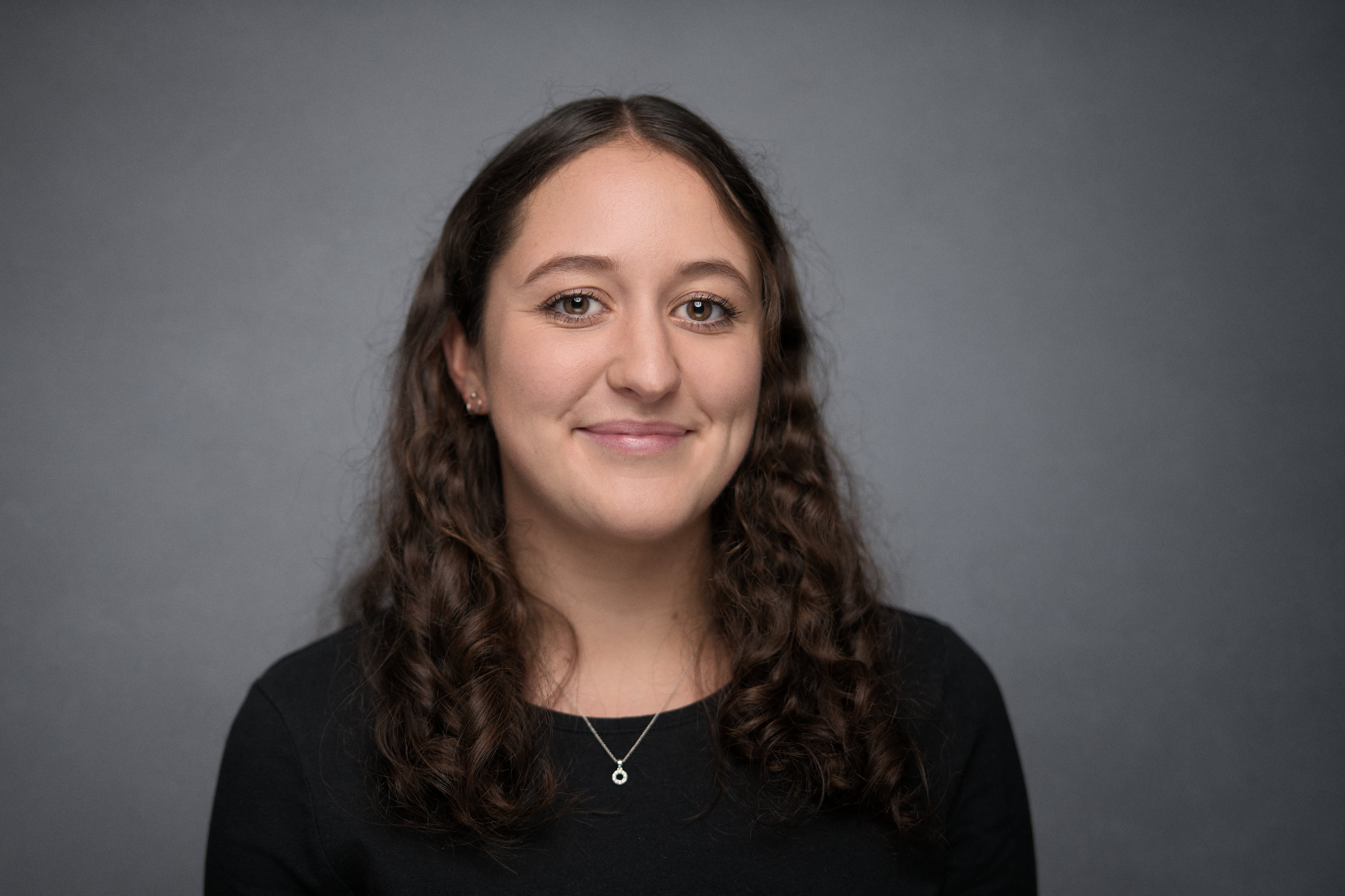Corporate headshot of a woman with long curly hair, smiling naturally. She is wearing a black top against a soft grey background, creating a professional and approachable look. The image captures subtle studio lighting emphasizing her features, contributing to an authentic expression. Photographed by Adam Toth of Tadam Photography in London, known for refined and elegant portrait photography.