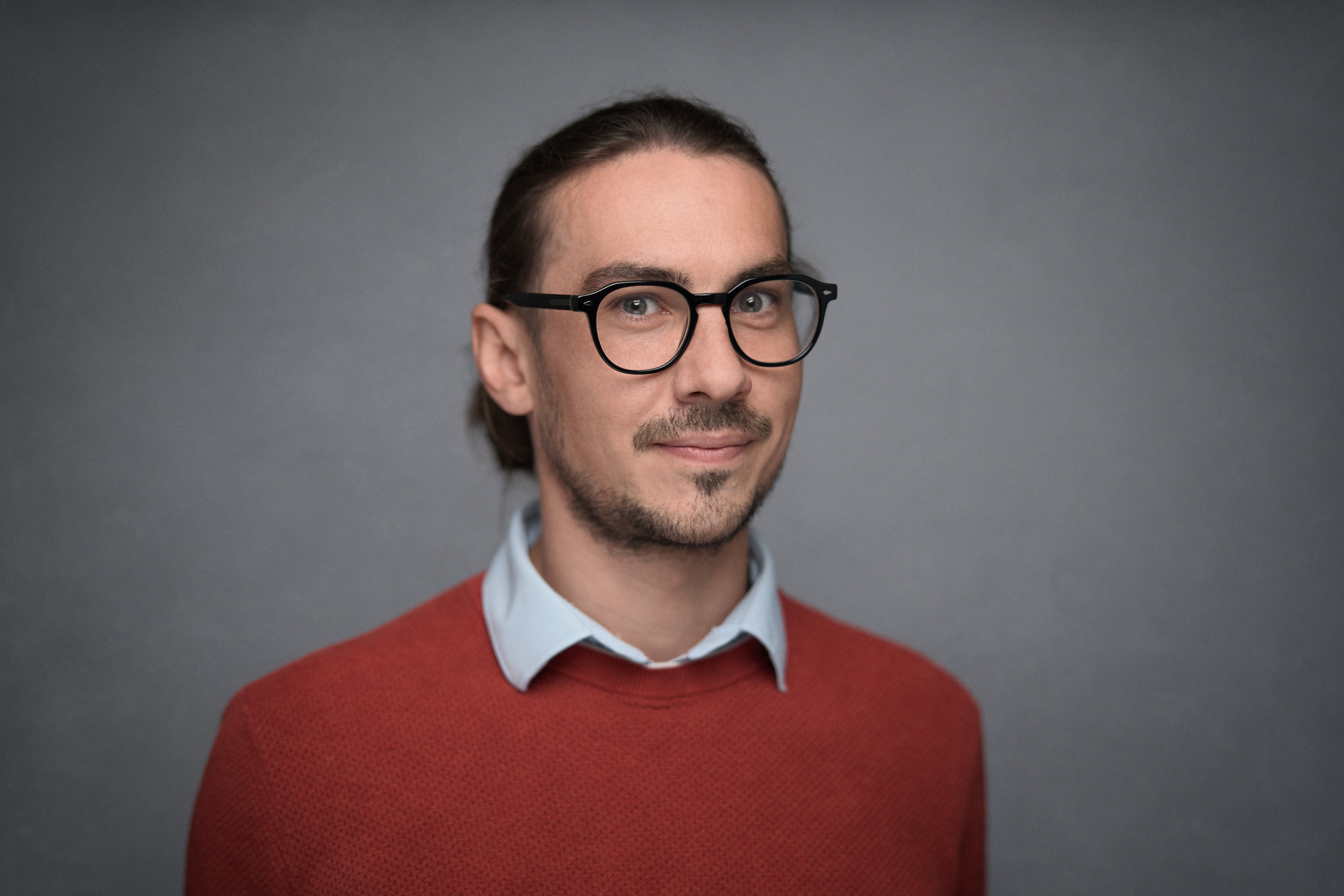 Corporate headshot by Adam Toth at Tadam Photography, featuring a man in a stylish red sweater and light blue shirt, exuding a confident yet approachable demeanor. Captured using professional studio lighting with a neutral gray background, this portrait reflects clarity and authenticity. Photographed in London, known for its dynamic business environment, showcasing the subject's engaging presence