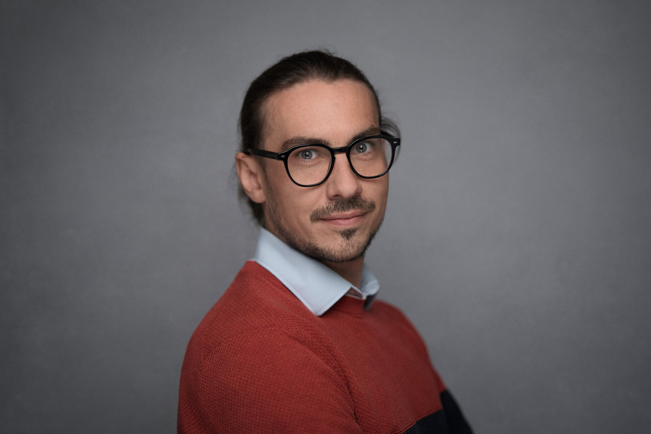 Man in a rust jumper and round glasses photographed over his shoulder, hair tied back, grey backdrop
