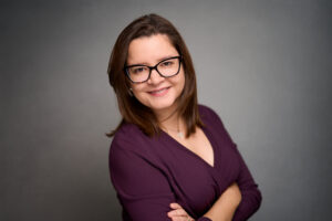 Woman with shoulder-length brown hair and black glasses wearing purple top, smiling arms crossed.