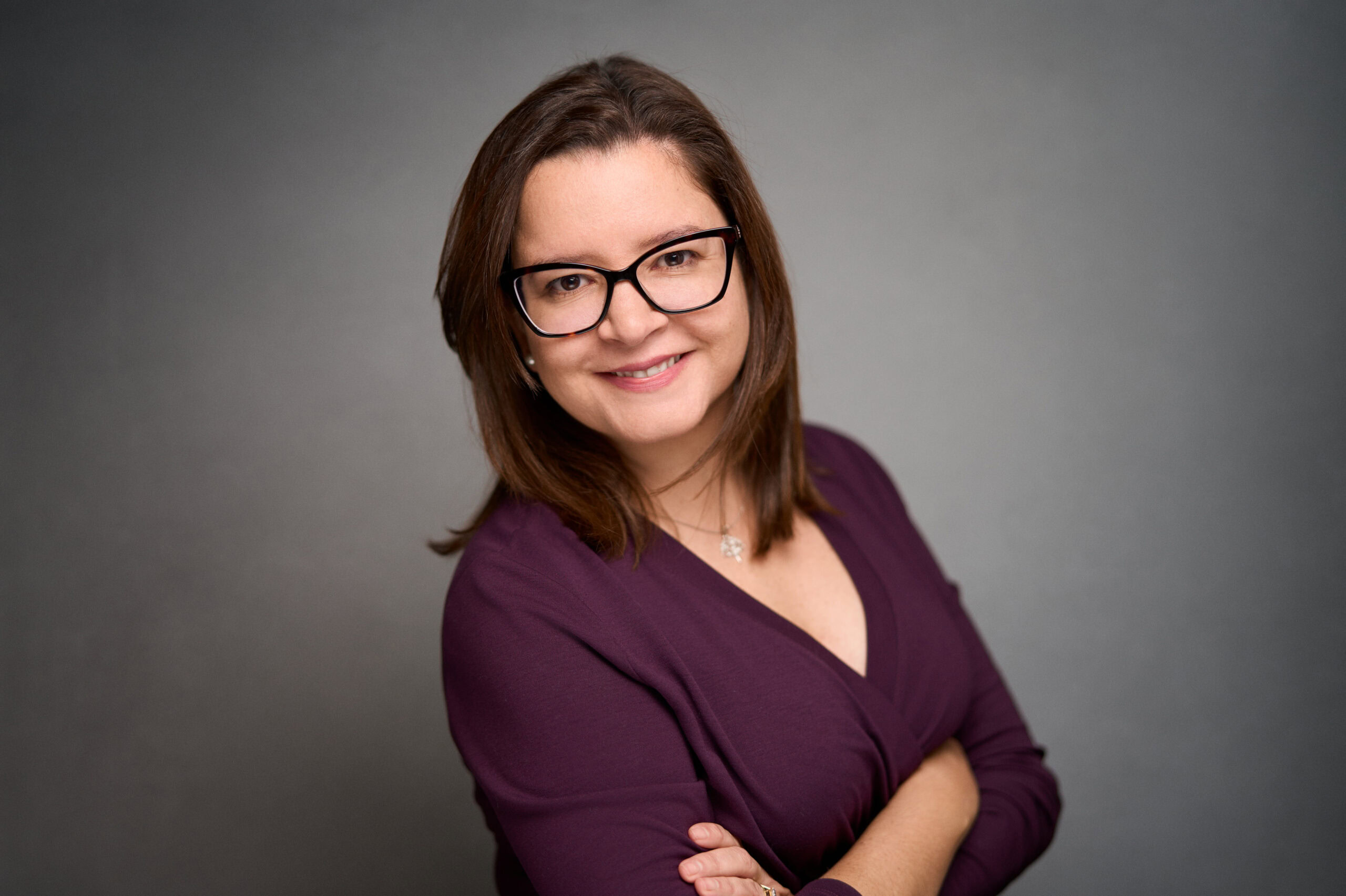 Woman with shoulder-length brown hair and black glasses wearing purple top, smiling arms crossed.
