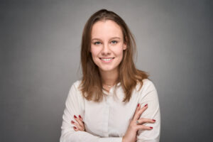 Young woman with long light-brown hair in a white blouse, arms crossed with red nails, smiling