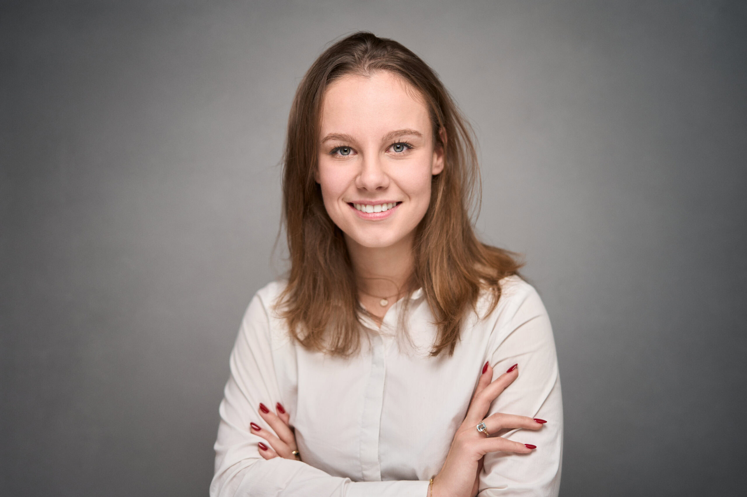 Young woman with long light-brown hair in a white blouse, arms crossed with red nails, smiling
