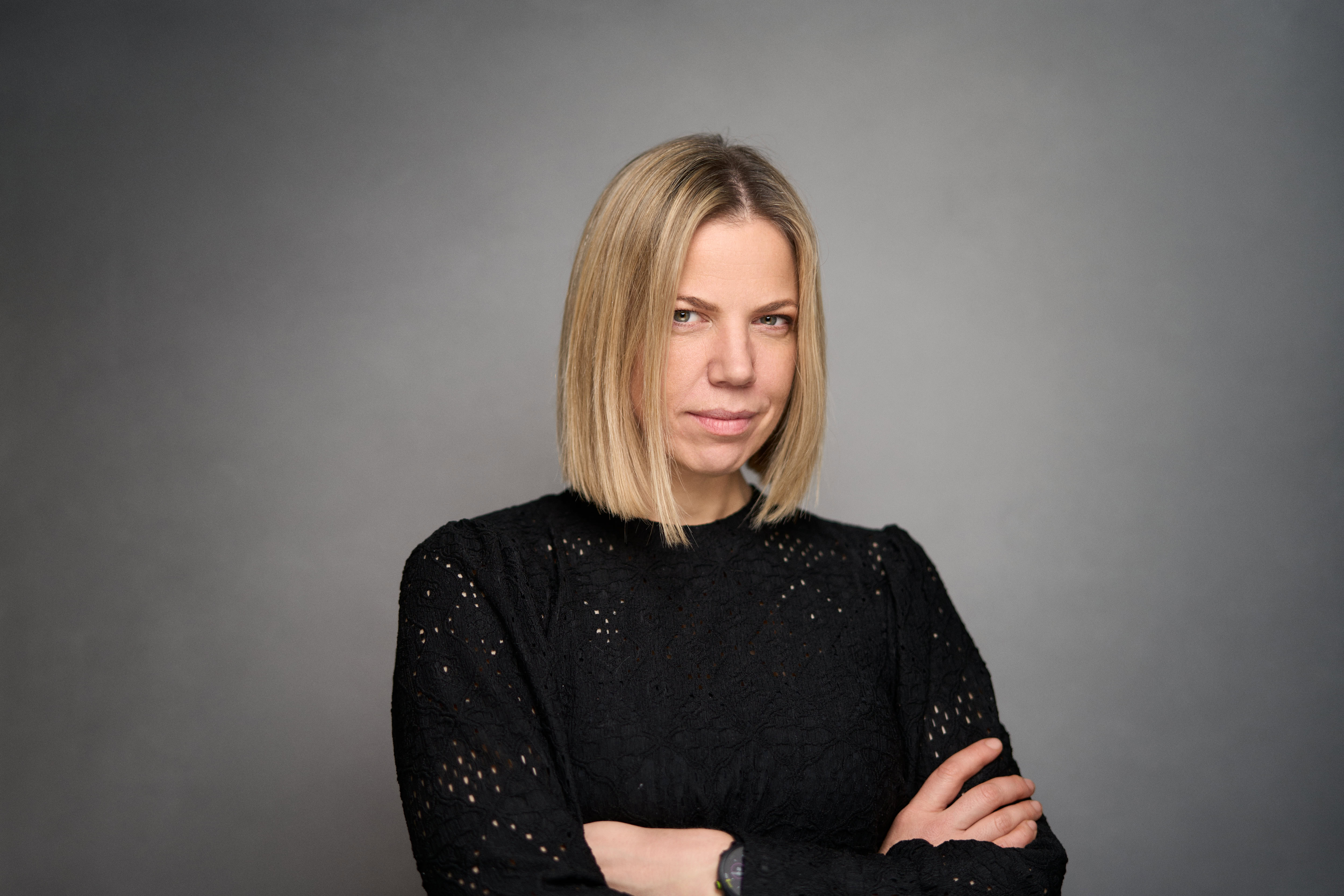 Corporate headshot by Adam Toth at Tadam Photography, featuring a confident professional in a black lace top, posing with arms crossed. Captured with professional studio lighting, the clean gray background enhances focus on the subject's natural, assured expression. Photographed in London, this image showcases modern business portraiture.