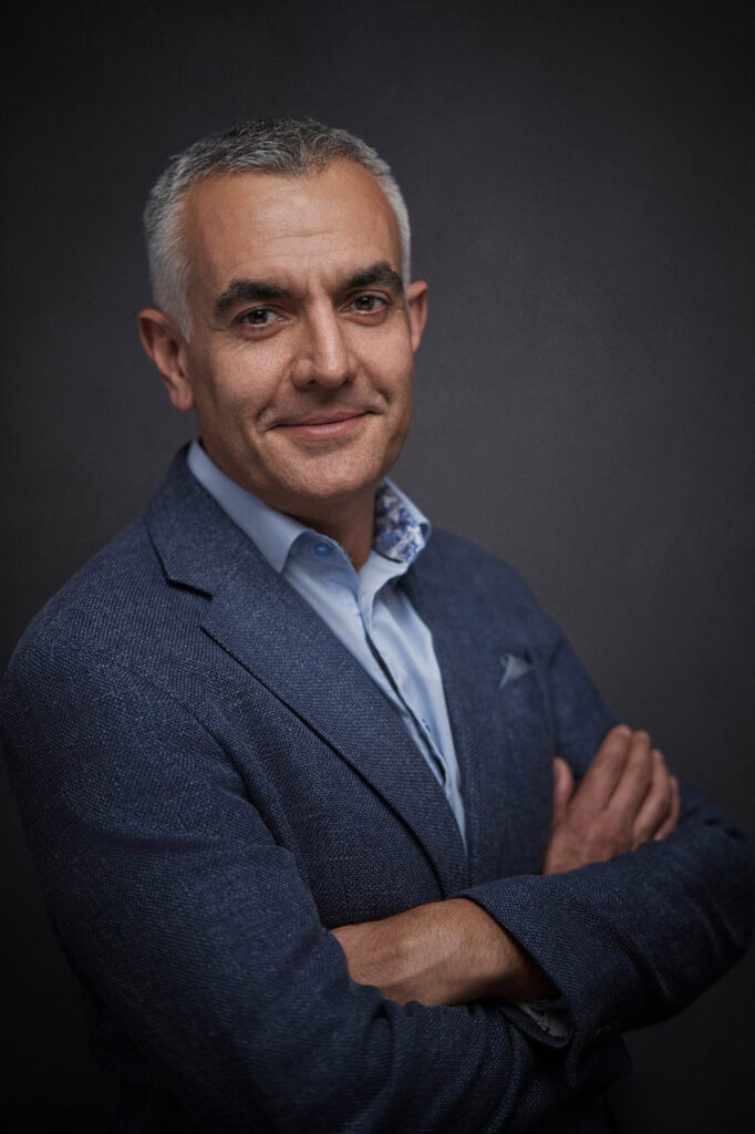 Nikhil in a blue blazer over a pale checked shirt, arms crossed with a slight smile, dark backdrop