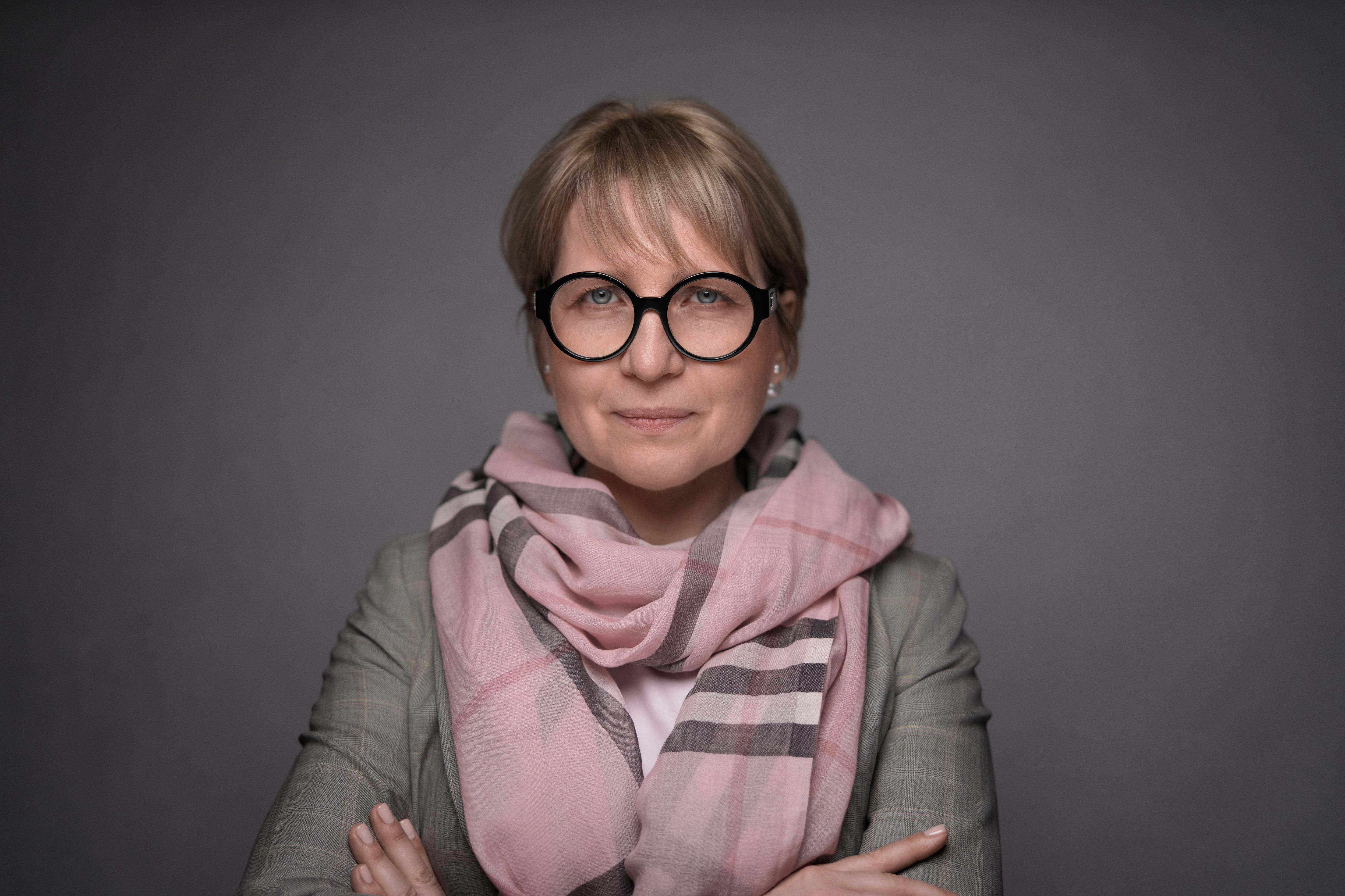 Corporate headshot featuring a professional woman wearing stylish glasses and a chic scarf, captured in a natural pose by Adam Toth of Tadam Photography. The image showcases professional studio lighting with a clean, muted background, emphasizing the subject's confident expression. This photograph represents the signature style of Tadam Photography, known for creating impactful corporate images in London, including