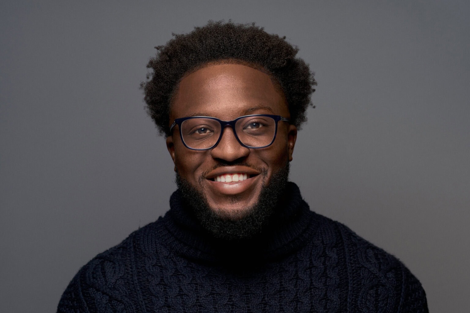 Smiling man with short afro hair and glasses in a navy cable-knit rollneck, on a grey studio backdrop
