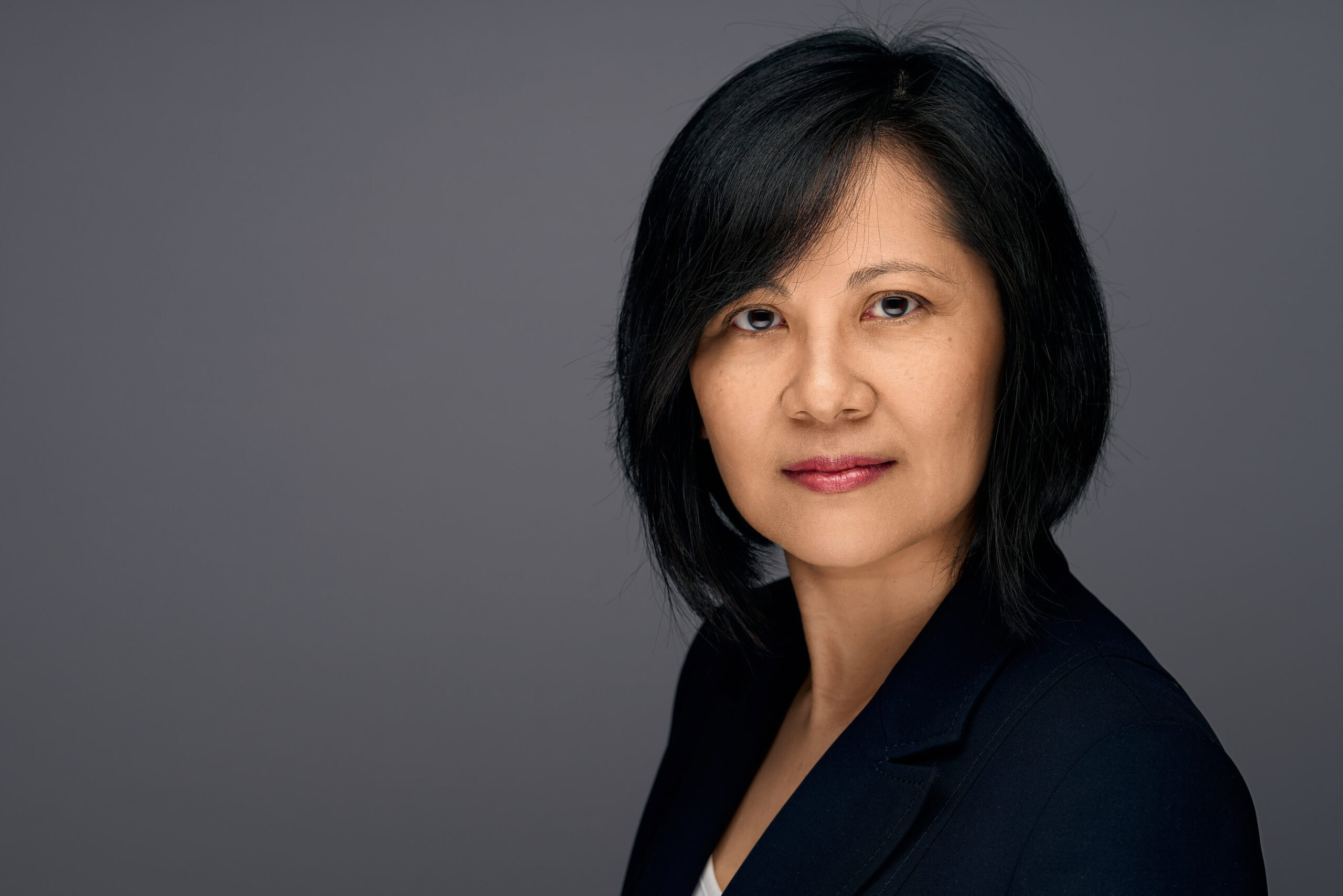 Woman with a sleek black bob in a navy blazer, poised expression on a soft grey backdrop