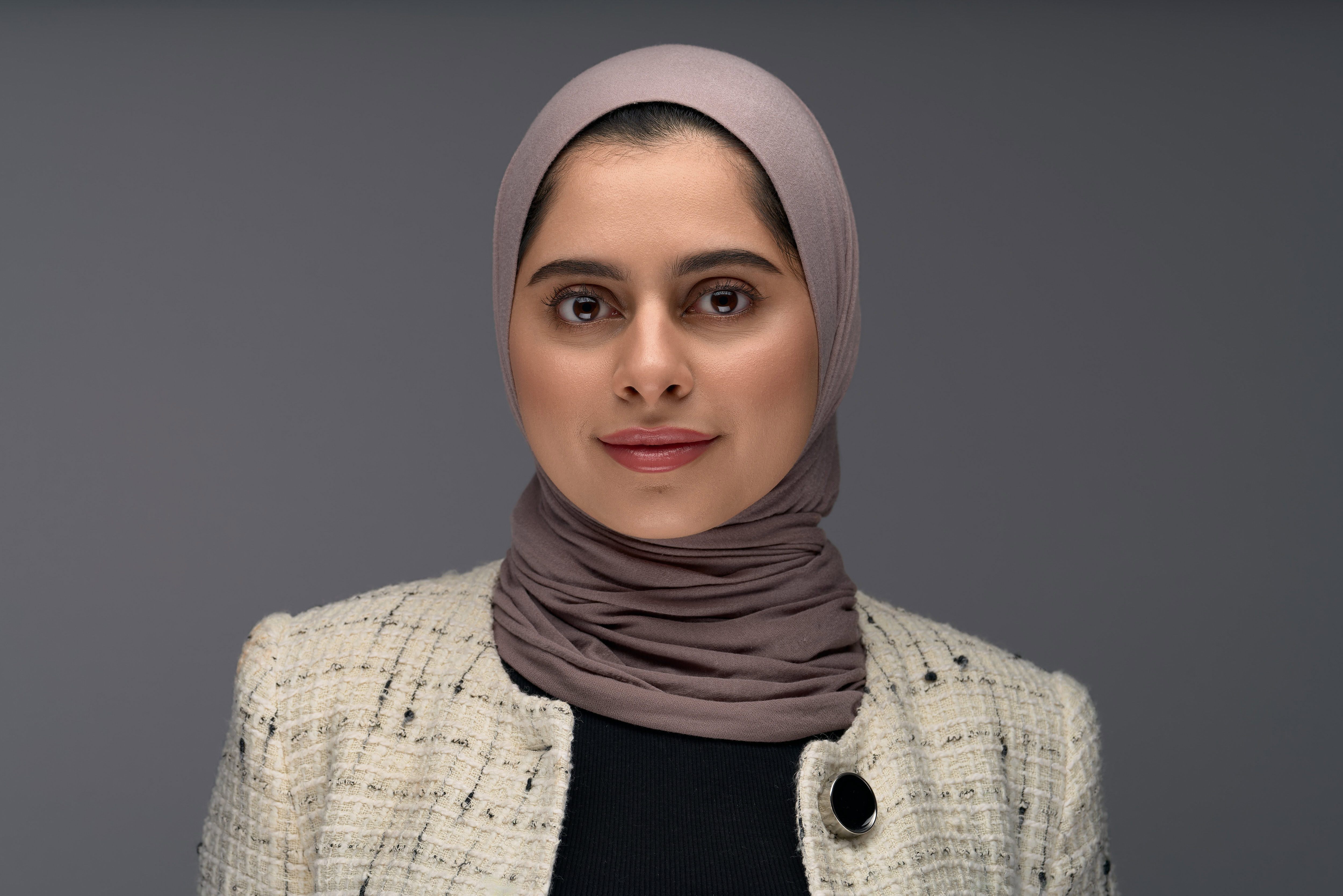 Corporate headshot featuring a confident professional woman in a textured beige jacket and taupe hijab, captured by Adam Toth of Tadam Photography. The individual presents a poised, natural pose against a neutral gray background, enhanced by professional studio lighting that emphasizes her engaging expression. Photographed in London, this image exemplifies the modern, polished style characteristic of