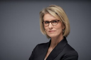 Middle-aged woman with short blonde hair, glasses, hoop earrings, and dark pinstripe blazer on gray background