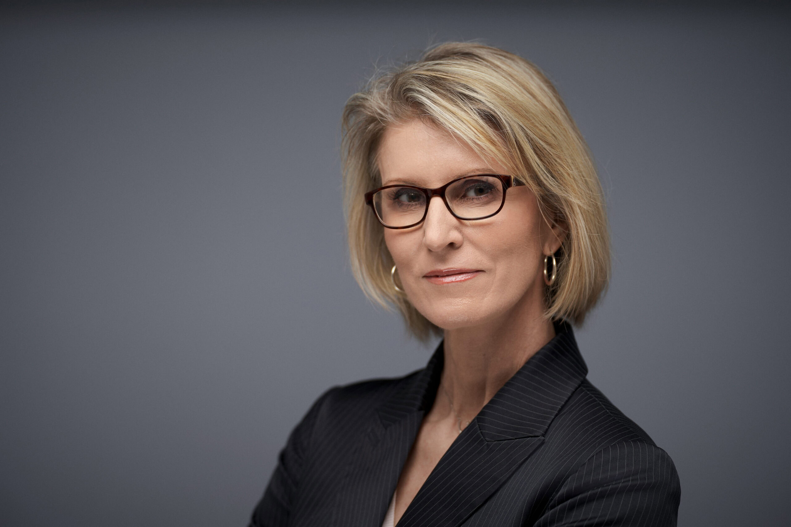 Middle-aged woman with short blonde hair, glasses, hoop earrings, and dark pinstripe blazer on gray background