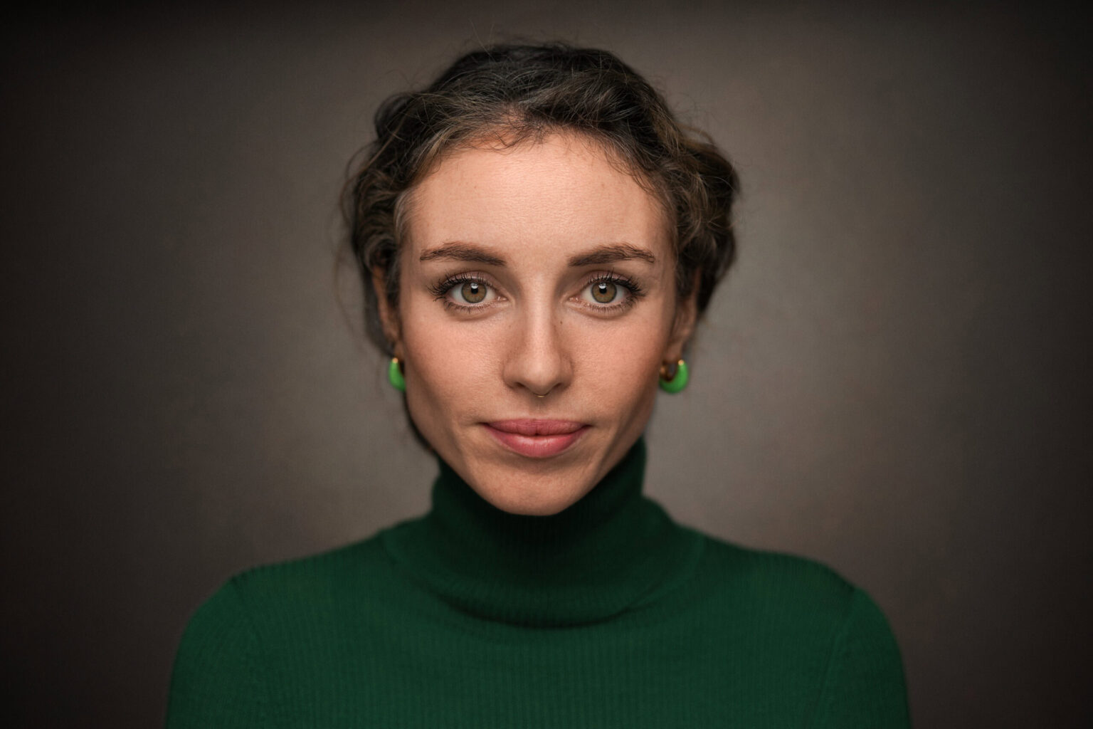Young woman with a braided updo in a green rollneck and green earrings, calm direct gaze on a grey backdrop